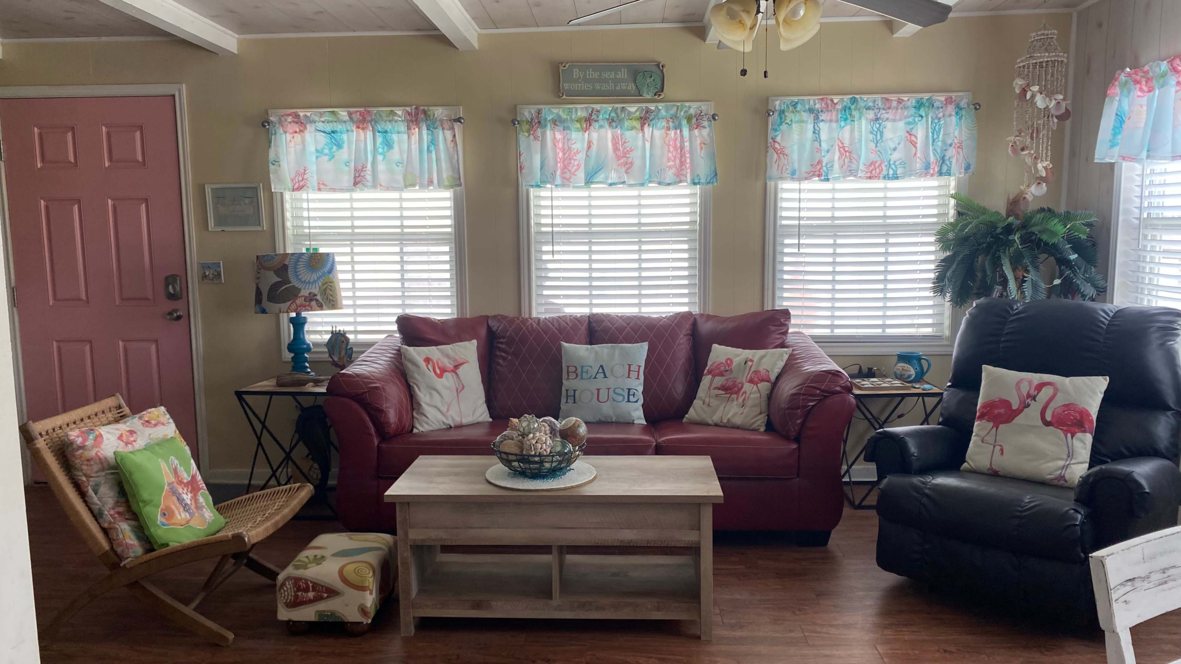 The living room features a red couch with flamingo-patterned pillows, a wooden coffee table, a reclining chair, and light blue window treatments.