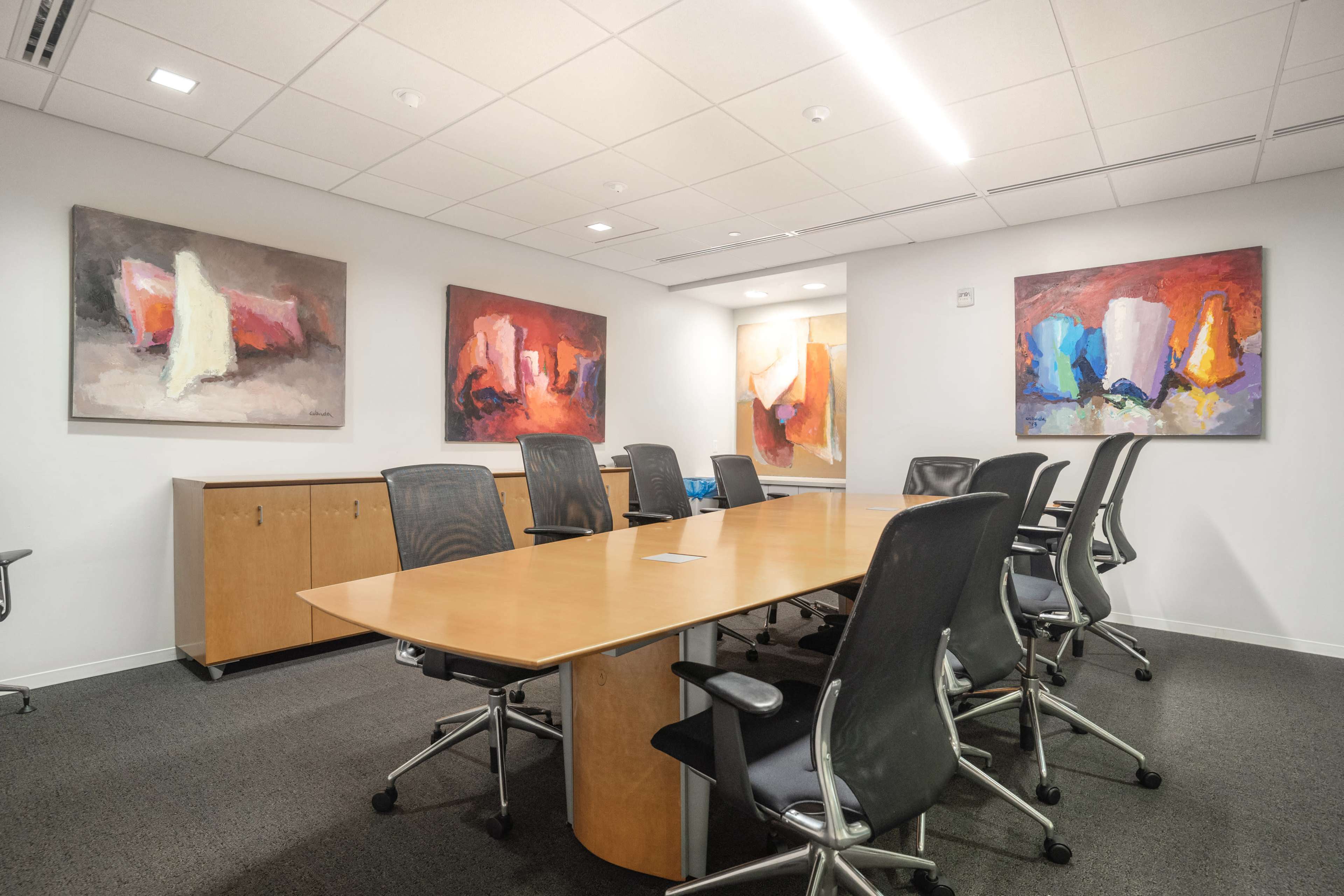 A modern conference room features a long wooden table surrounded by black chairs and colorful abstract paintings on the walls.