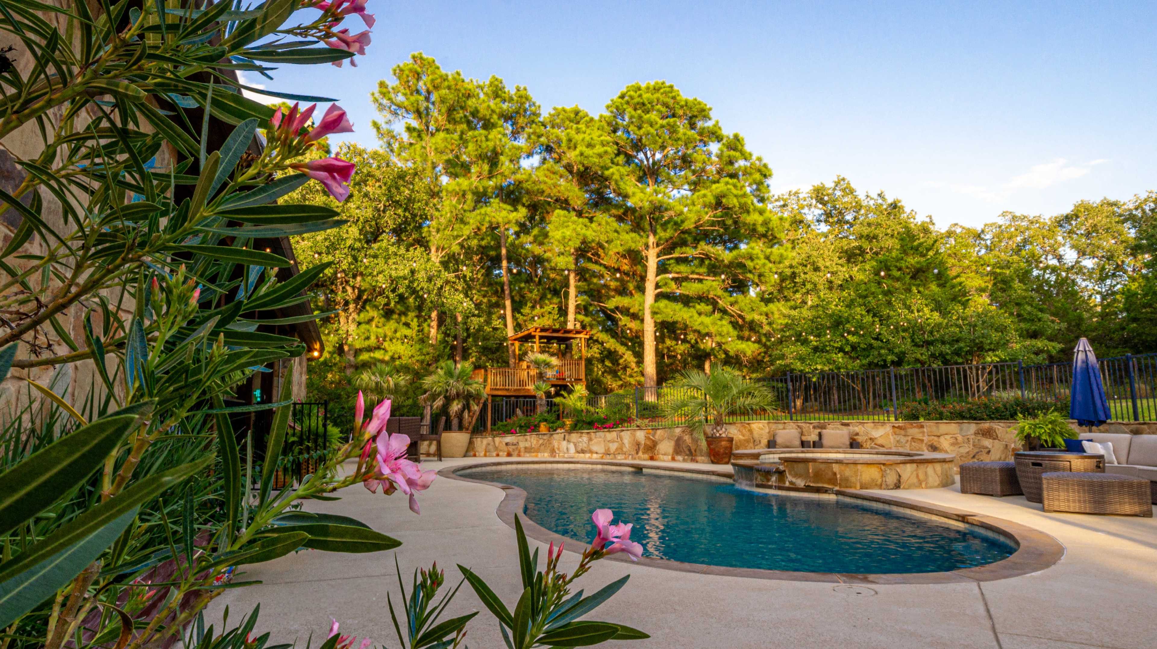 The image shows a curved swimming pool surrounded by greenery and a stone patio with outdoor seating and a play structure in the background.
