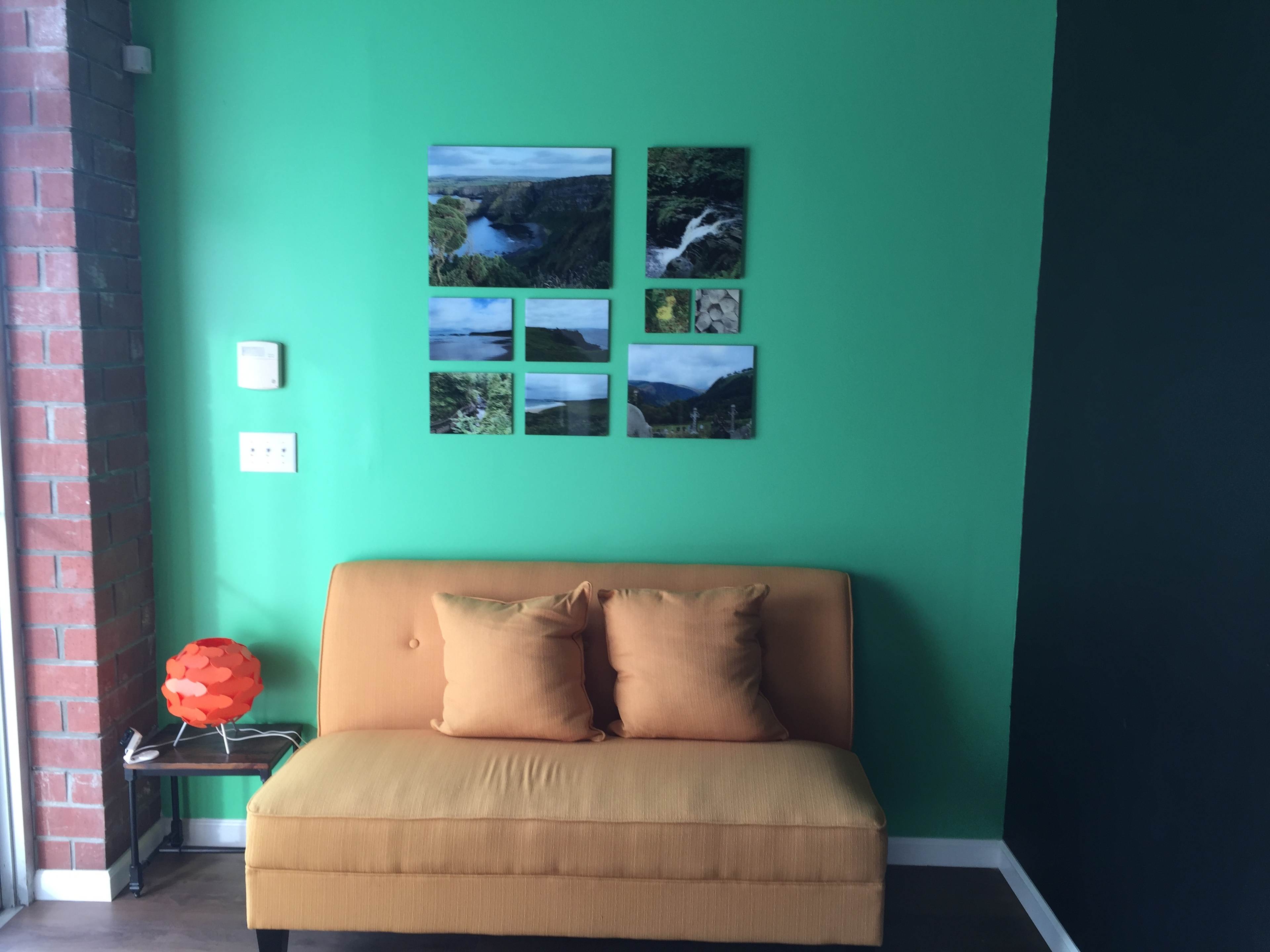 A small orange couch sits against a vibrant green wall, adorned with a series of landscape photographs above it, while a decorative lamp rests on a nearby side table.