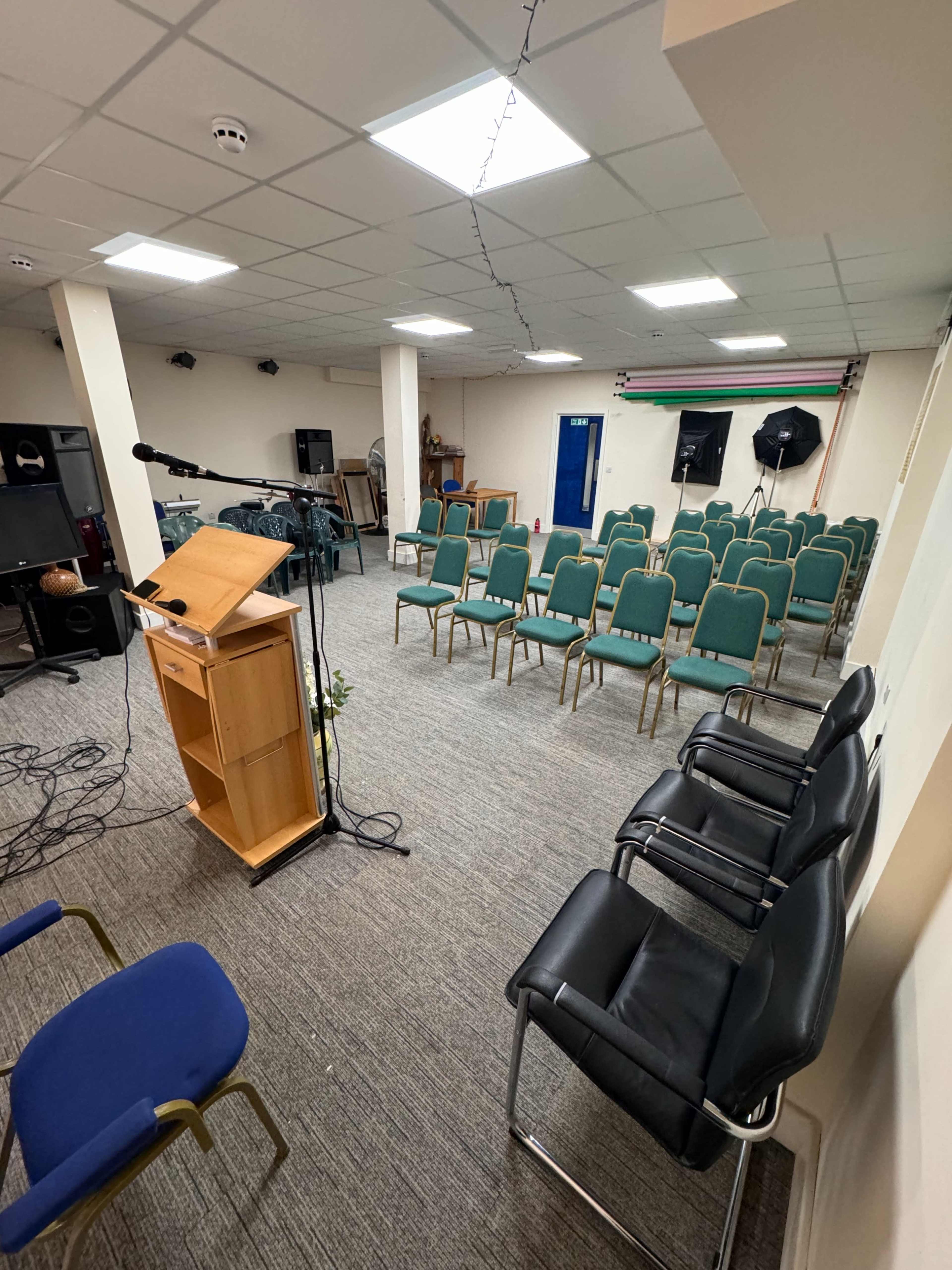 The image shows a conference room set up with rows of green chairs facing a wooden podium, with additional black chairs positioned along one side.