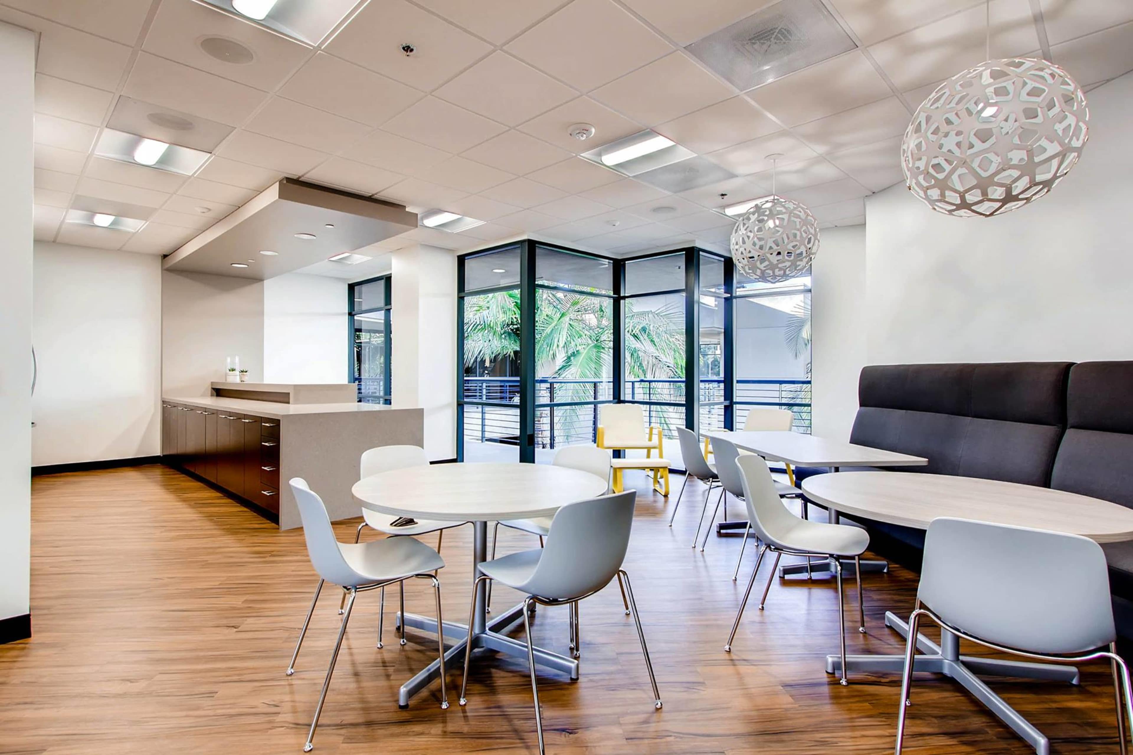 The image shows a modern break room with round tables, chairs, and large windows revealing greenery outside.