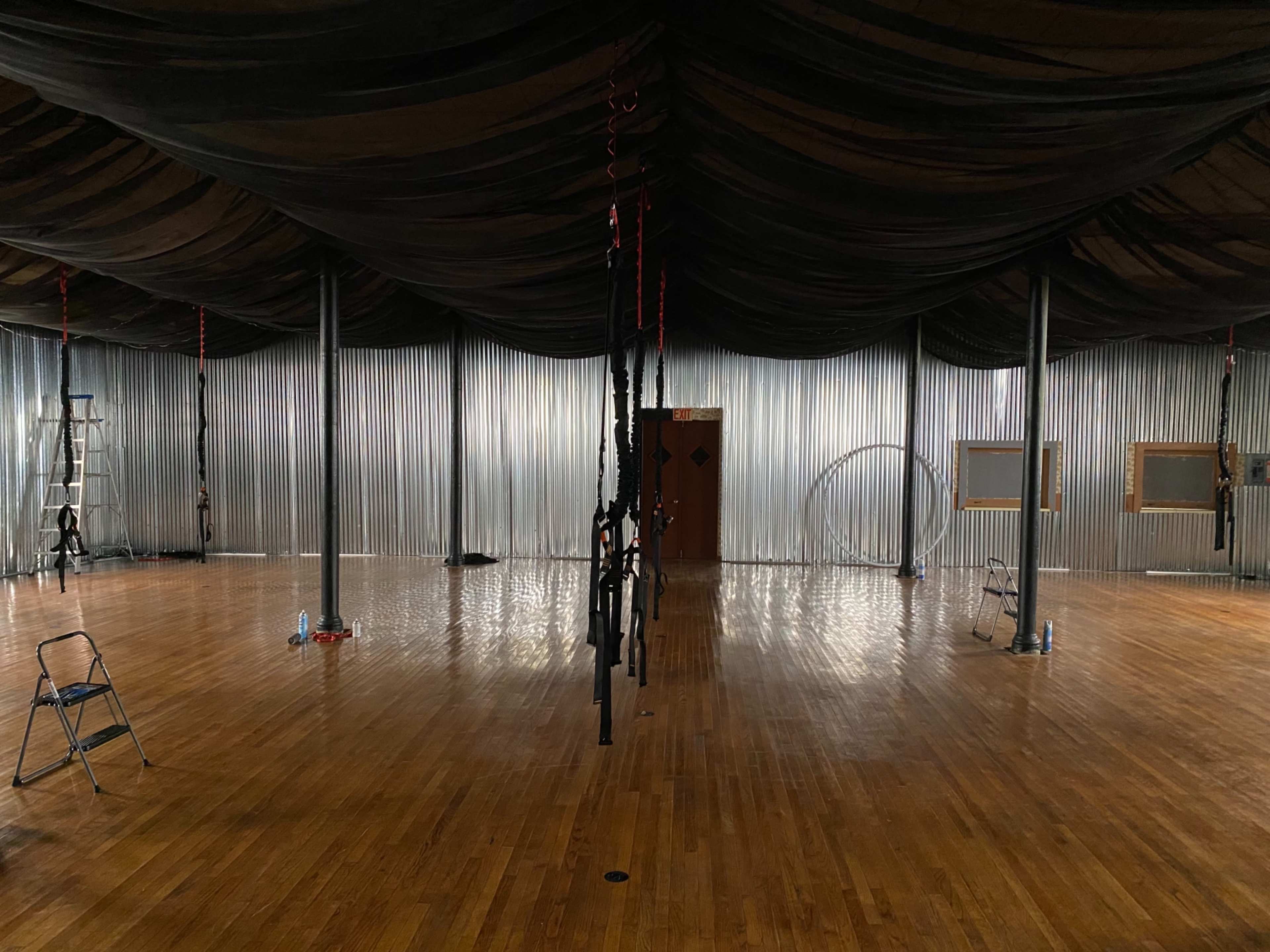 An empty indoor space with a polished wooden floor, black draped fabric overhead, and various aerial equipment suspended from the ceiling.