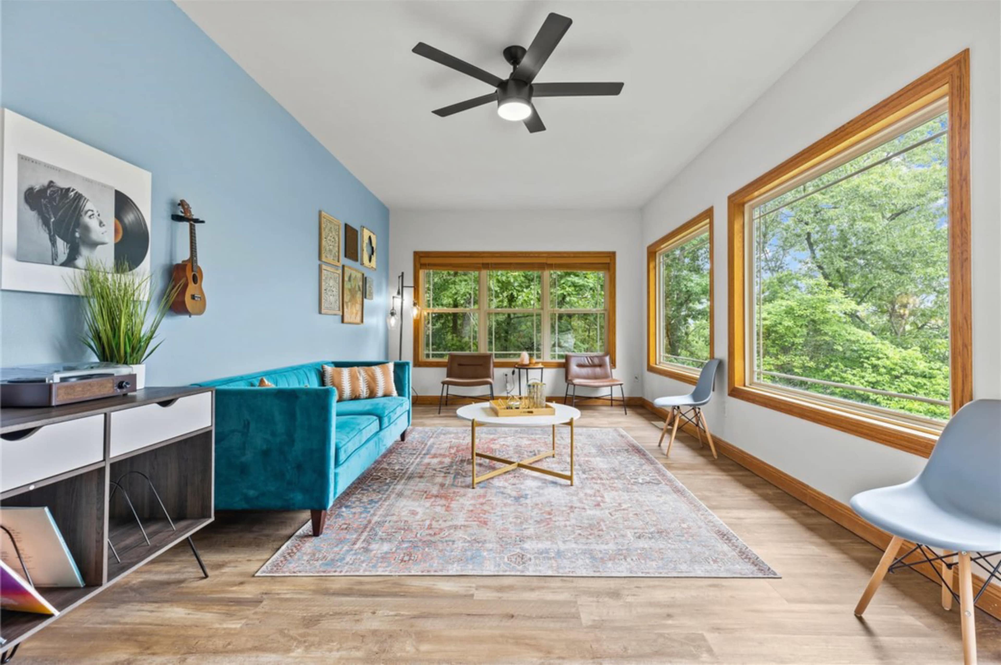 The room features a blue accent wall, large windows with views of greenery, and minimalist furniture including a turquoise couch and a coffee table on a patterned rug.