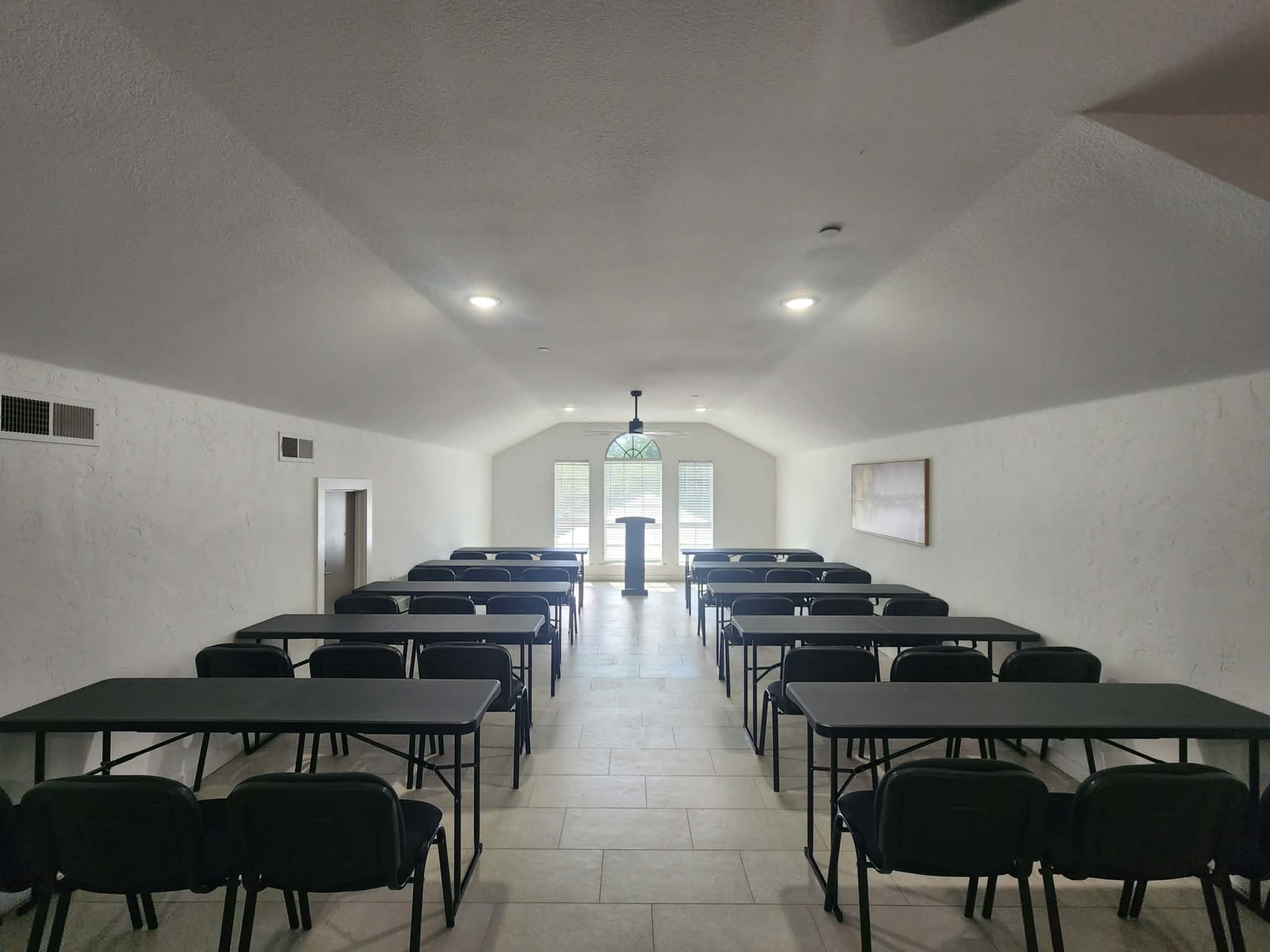A spacious meeting room features rows of black tables and chairs, with a podium at the front and large windows illuminating the space.