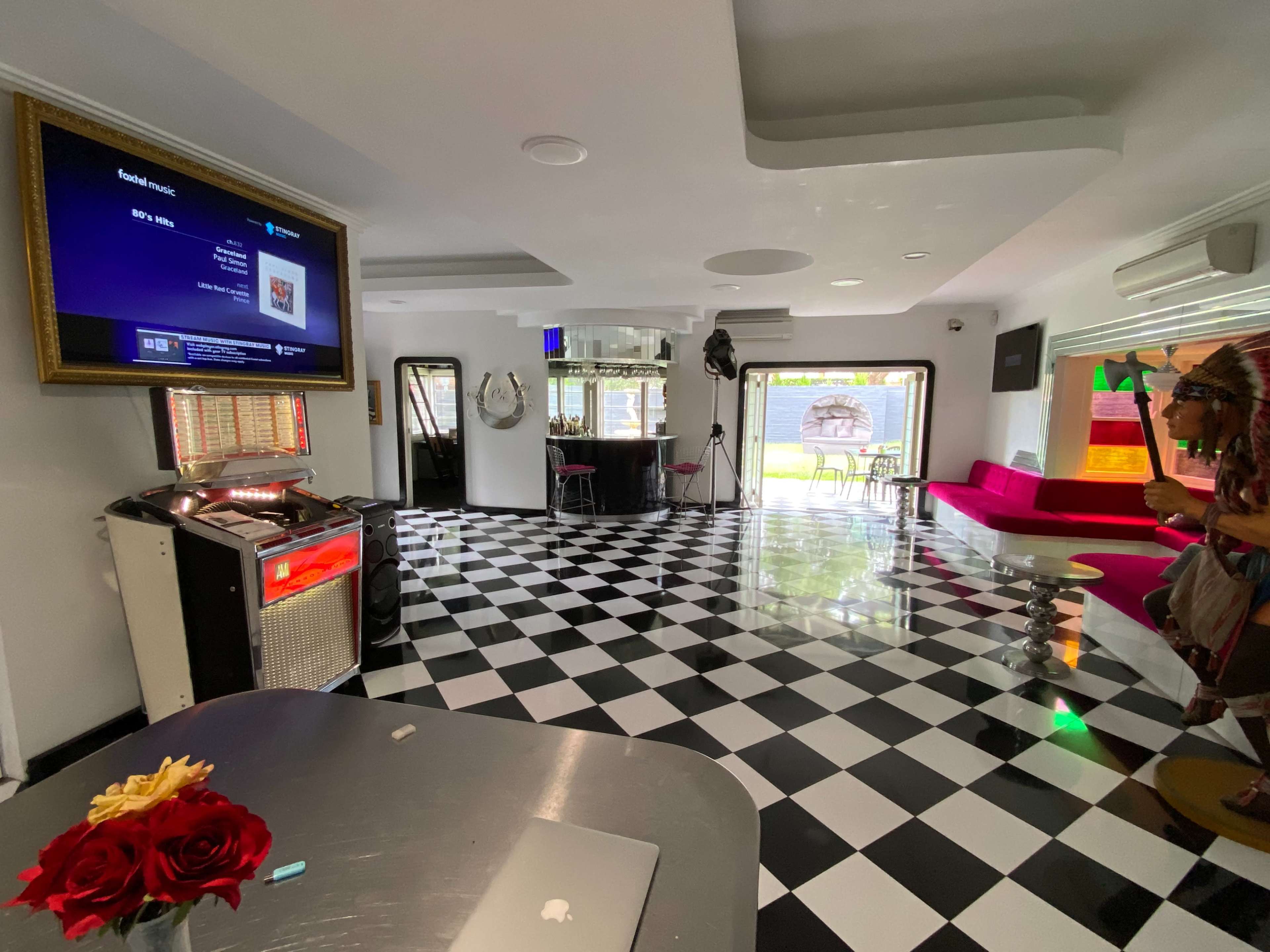 A modern room with checkered black and white flooring, a jukebox, and a red seating area, featuring large windows that open to a garden view.