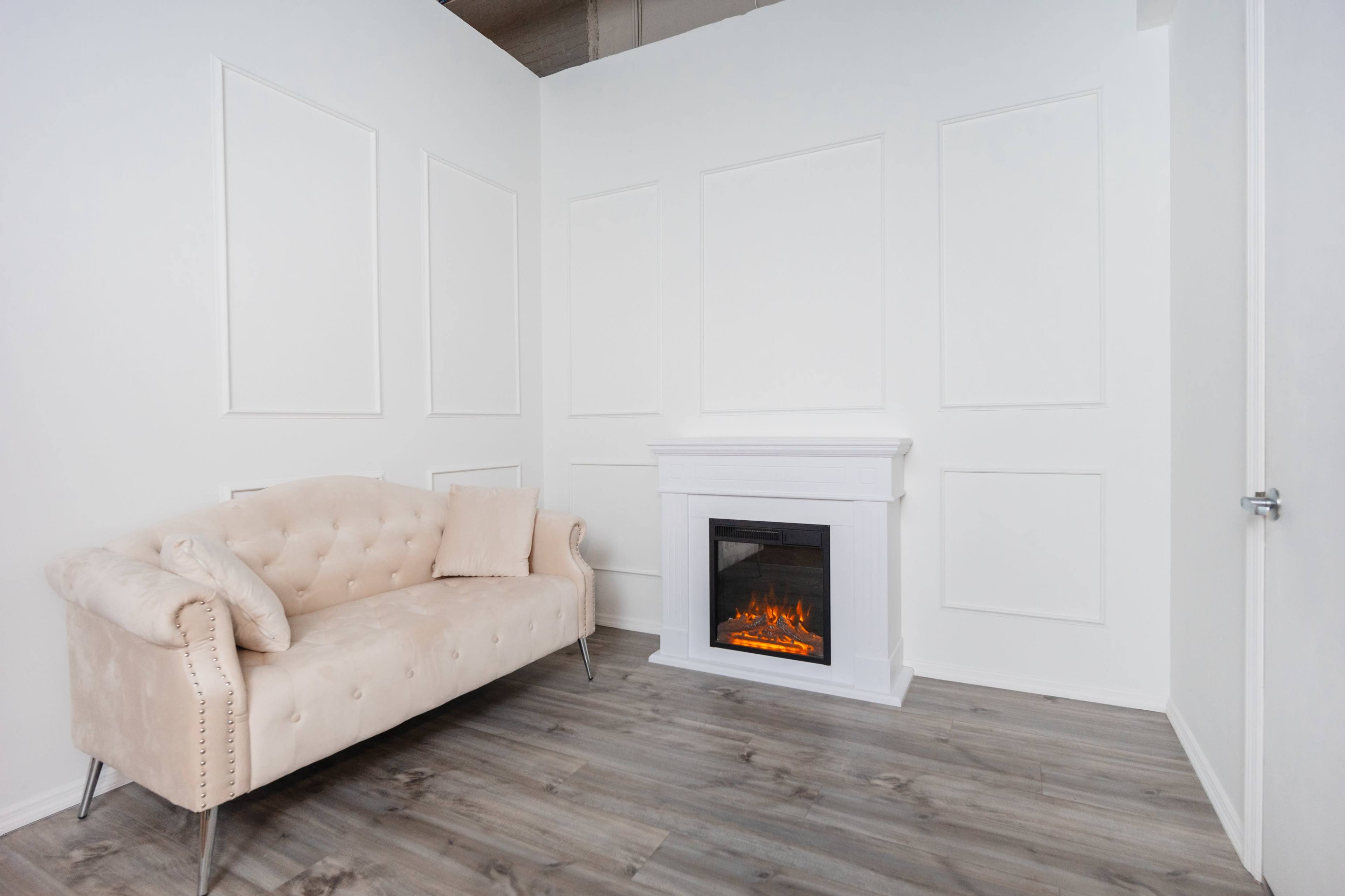 The image shows a minimalist room featuring a beige tufted sofa and an electric fireplace set into a white wall with recessed paneling.