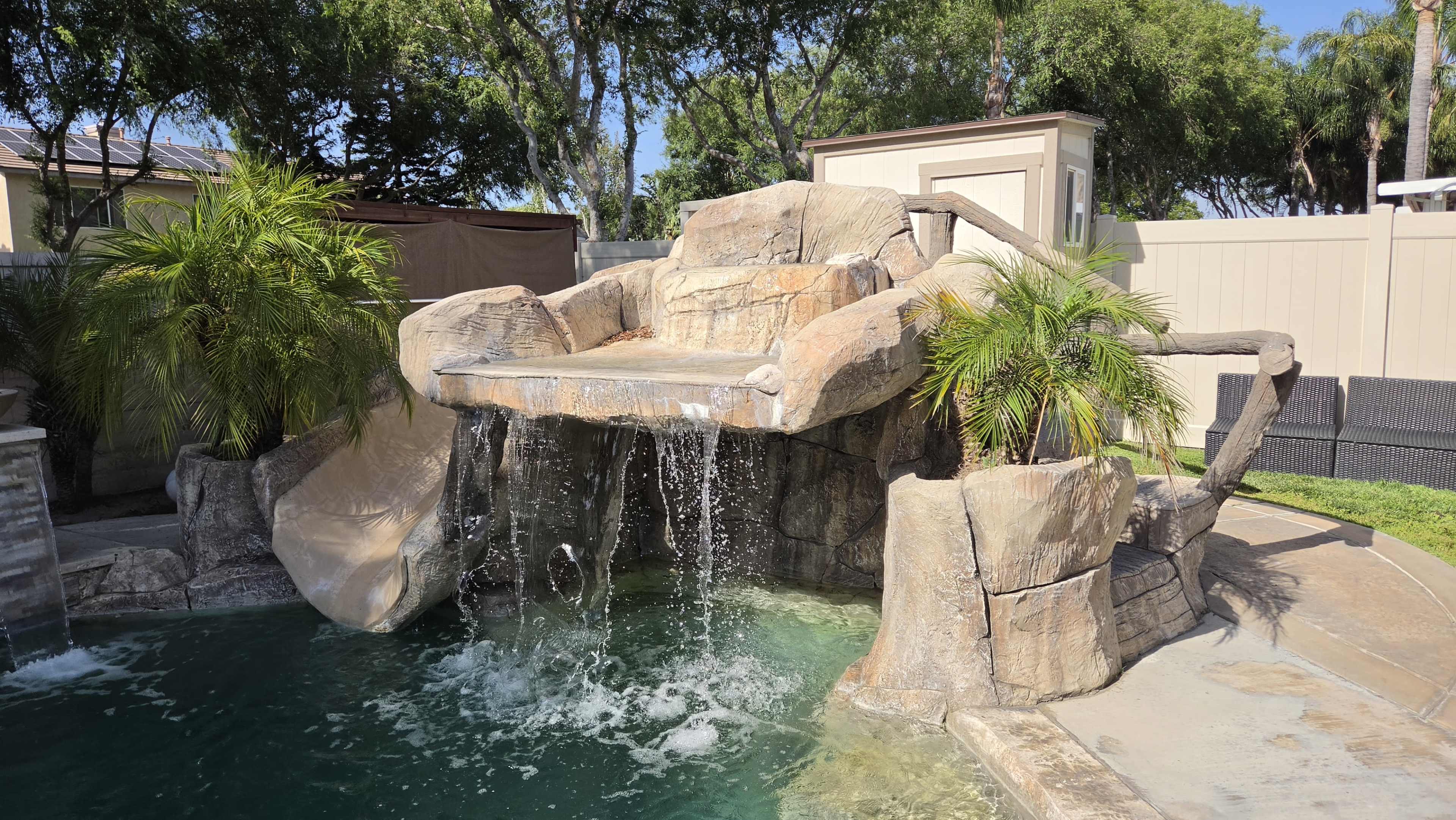 A rock formation with a waterfall cascades into a swimming pool, featuring tropical plants nearby.