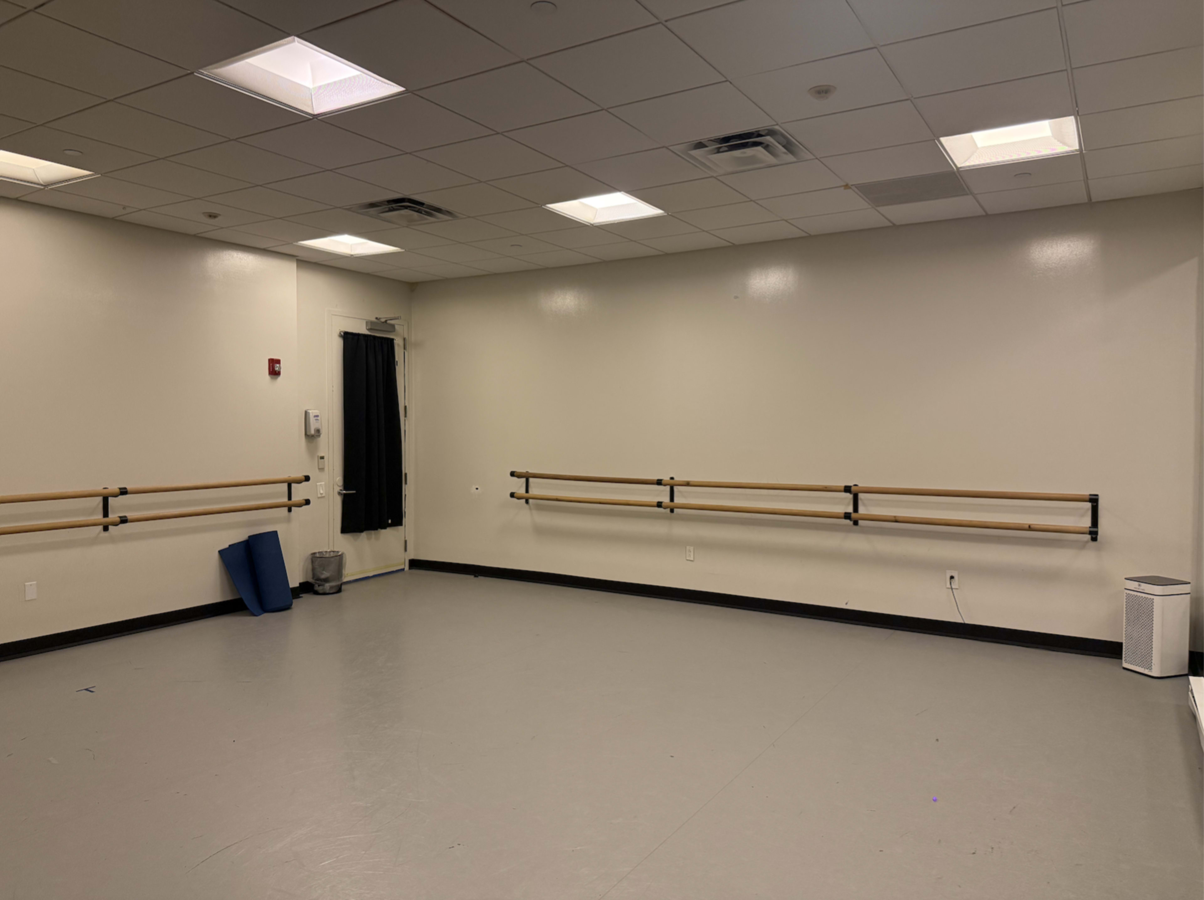 The image shows an empty dance studio with gray floors, wall-mounted ballet barres, and a door leading to the outside.