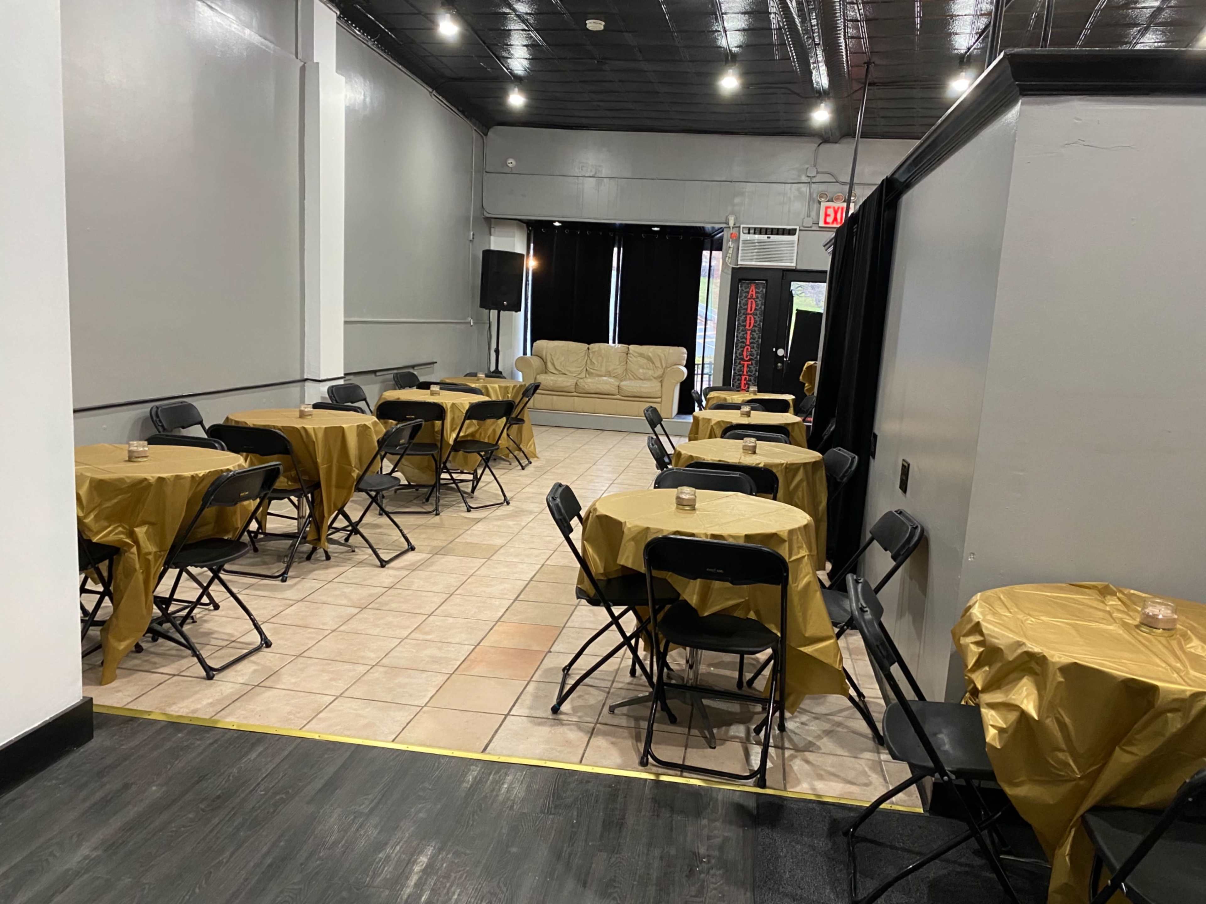 A spacious room set up with several round tables covered in gold tablecloths and black folding chairs, with a couch in the background.