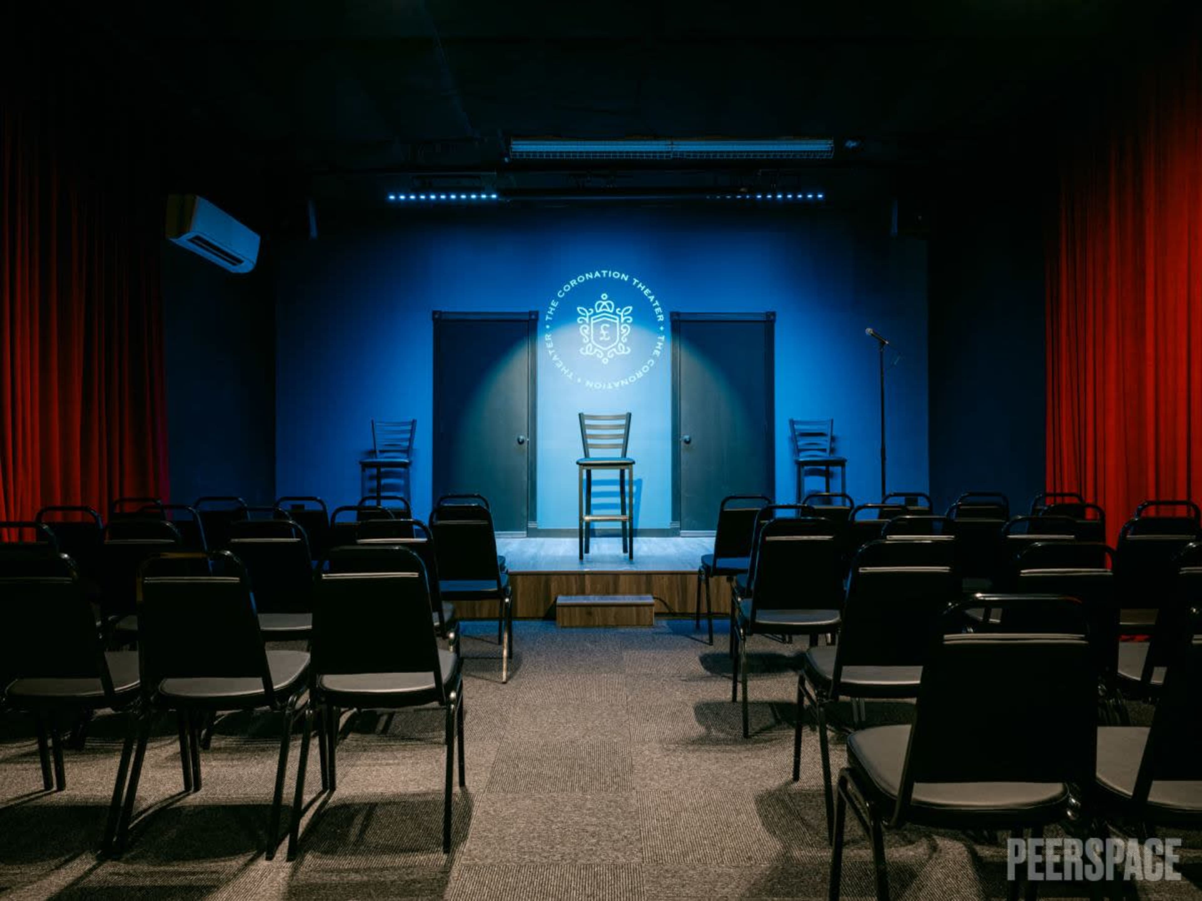 A dimly lit performance space with several rows of empty black chairs facing a simple stage featuring a logo, a microphone stand, and two closed doors.