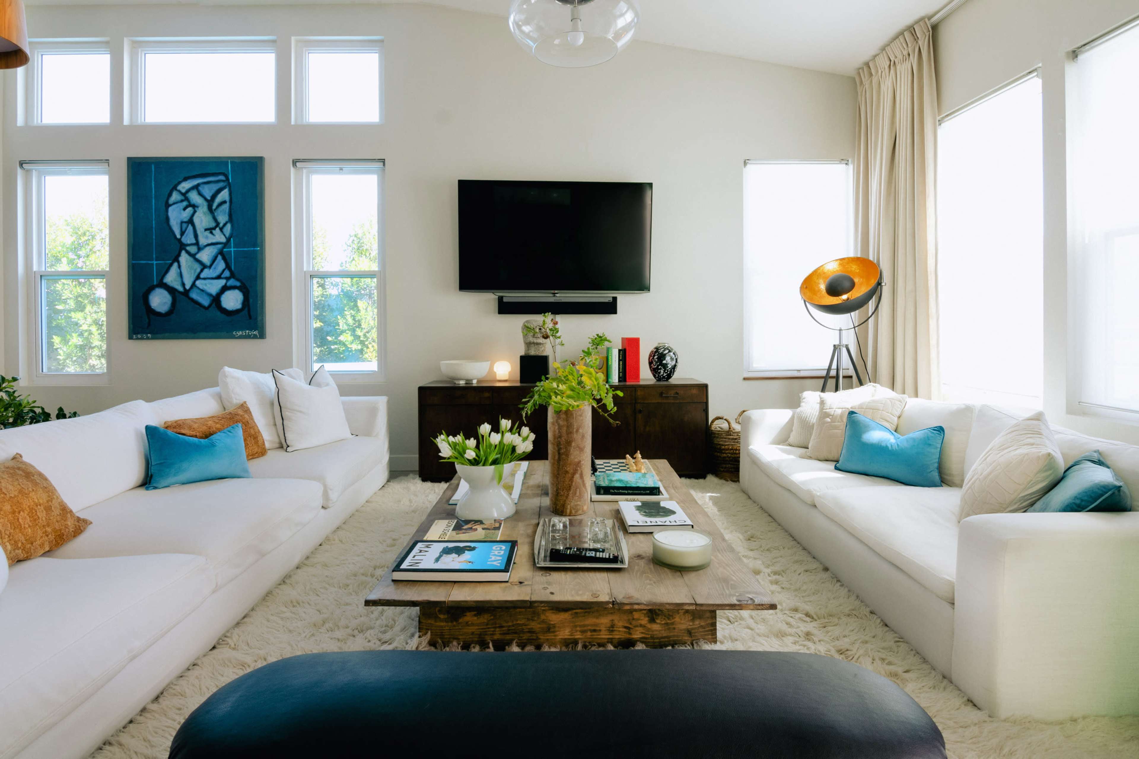 A spacious living room features white sofas, a wooden coffee table, a large TV on the wall, and framed artwork beside large windows.