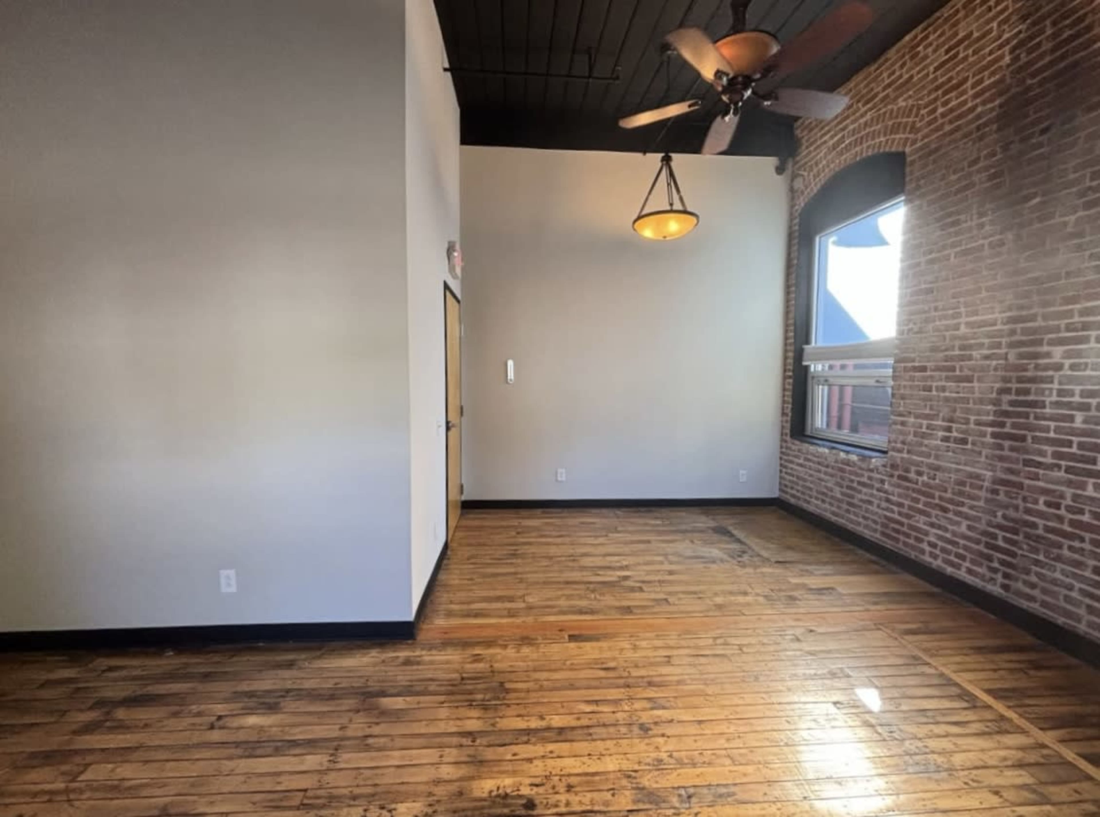 The image shows an empty room with exposed brick walls, wooden flooring, and a ceiling fan.