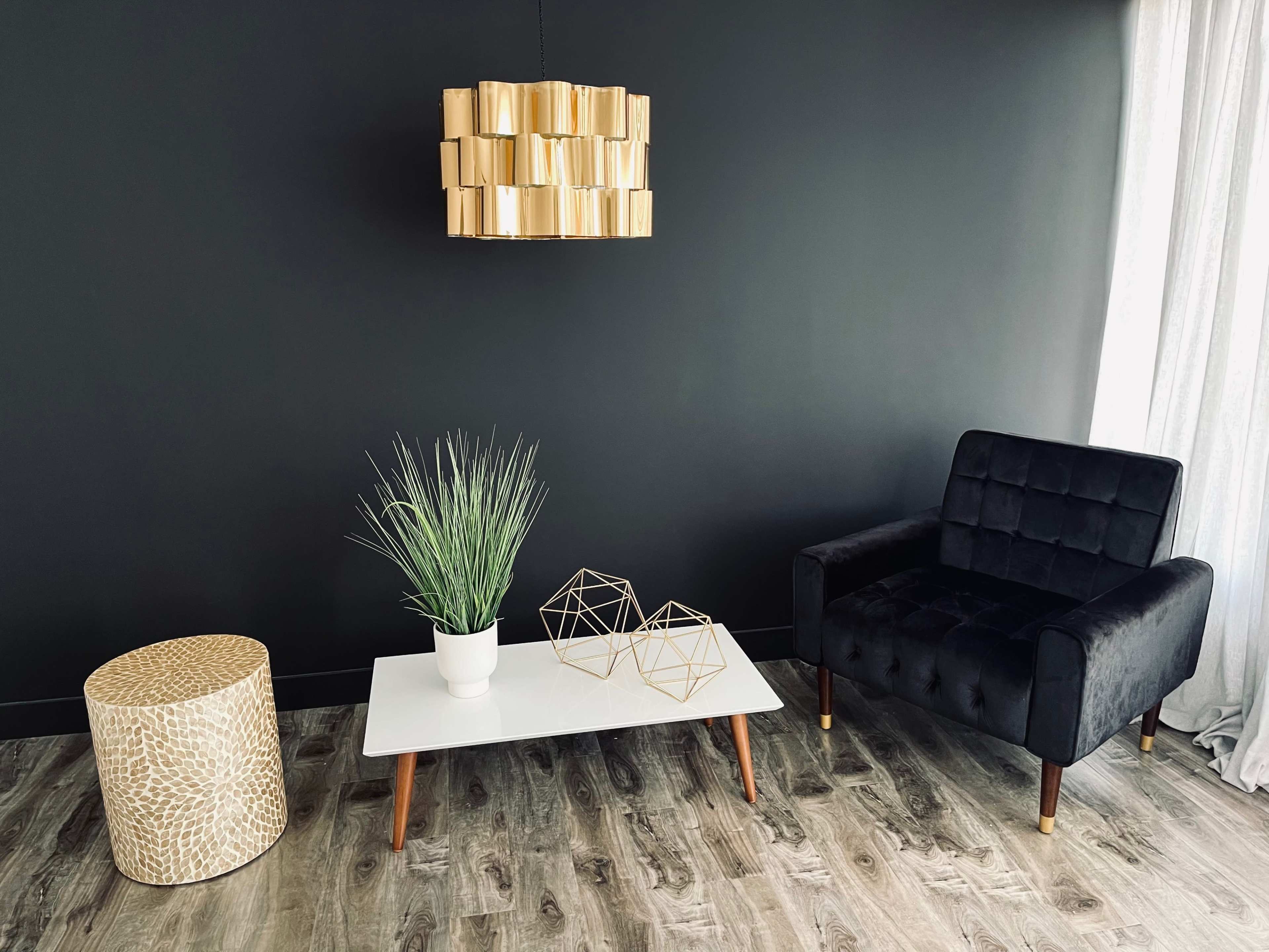 A modern living room with a black wall, a gold pendant light, a black upholstered chair, a white coffee table with geometric decorations, a potted plant, and a gold-patterned stool.