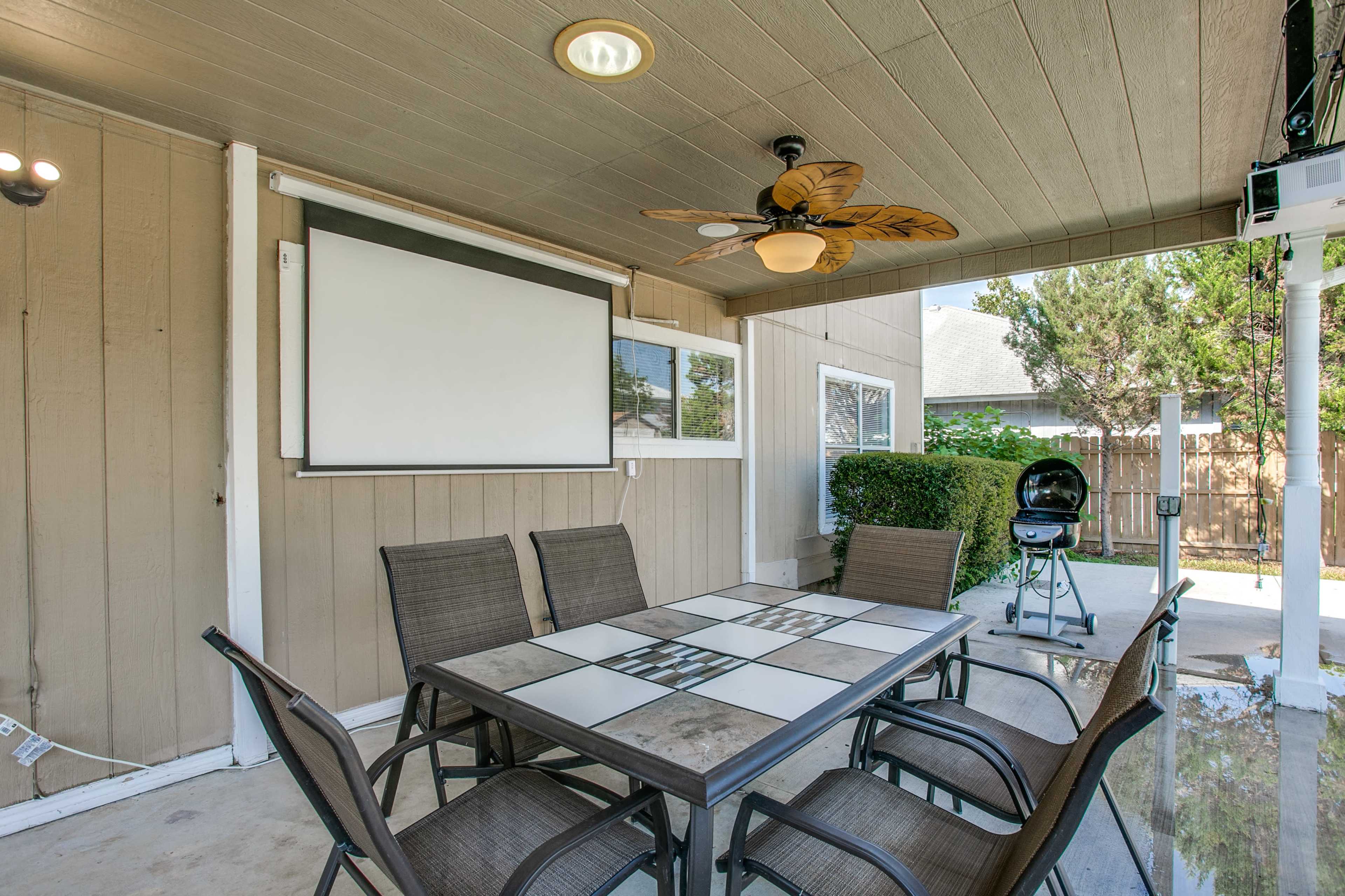A covered patio area with a dining table, six chairs, a ceiling fan, a movie screen, and a charcoal grill in the background.