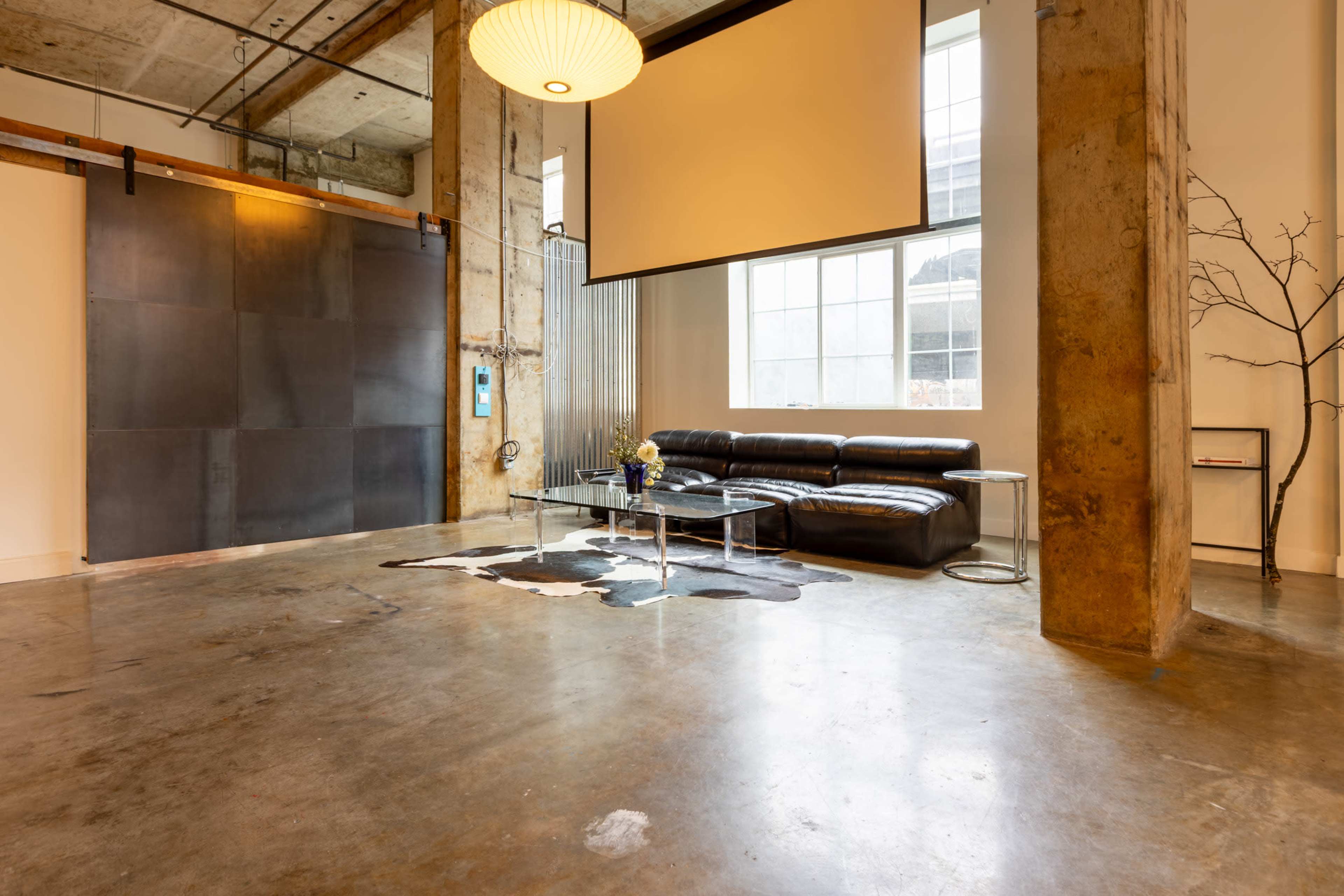 The image shows a spacious, modern living area featuring a dark sectional sofa, a glass coffee table, and large windows with natural light.