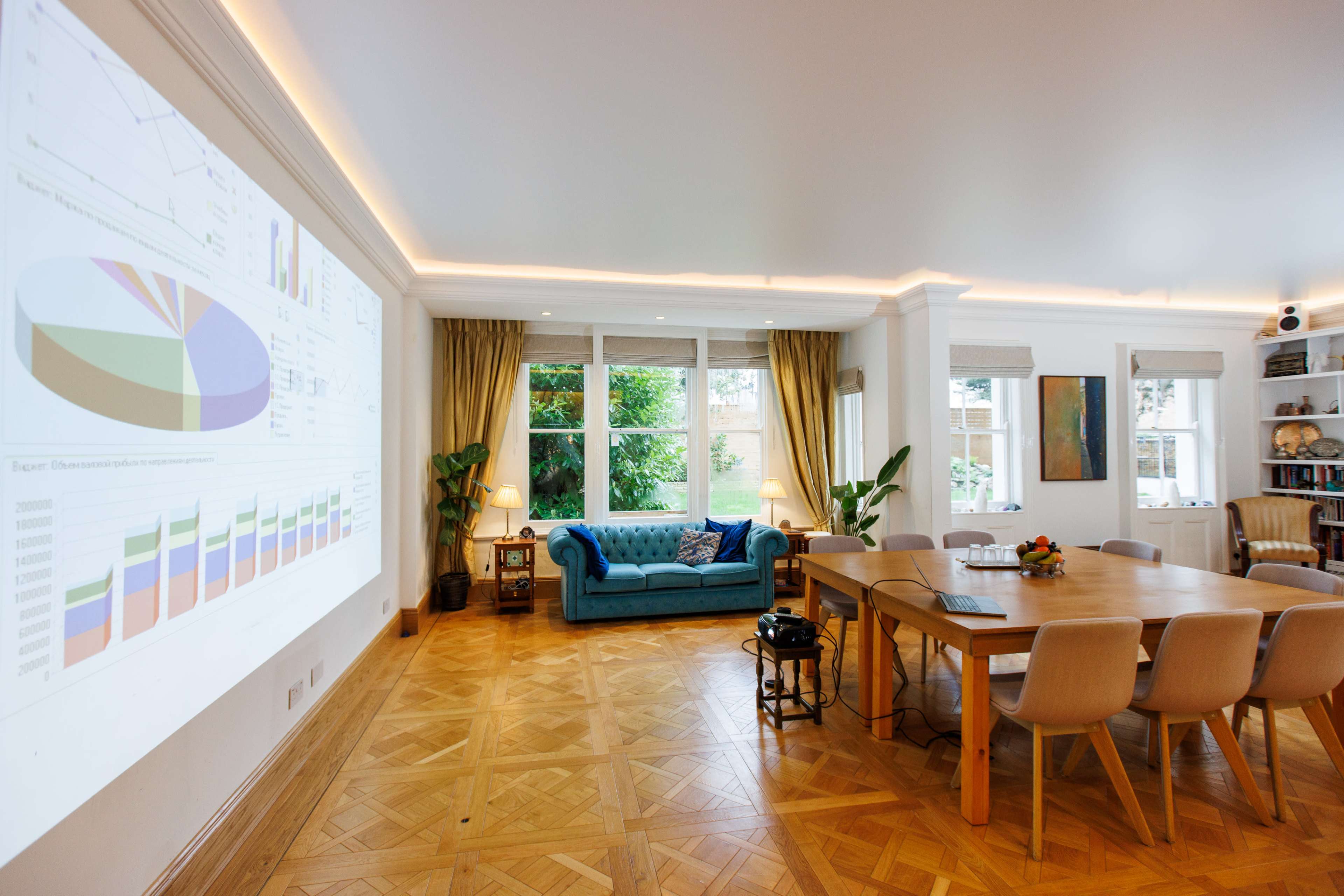 A cozy room features a turquoise sofa, a wooden dining table surrounded by chairs, and a large projection of colorful charts on the wall.