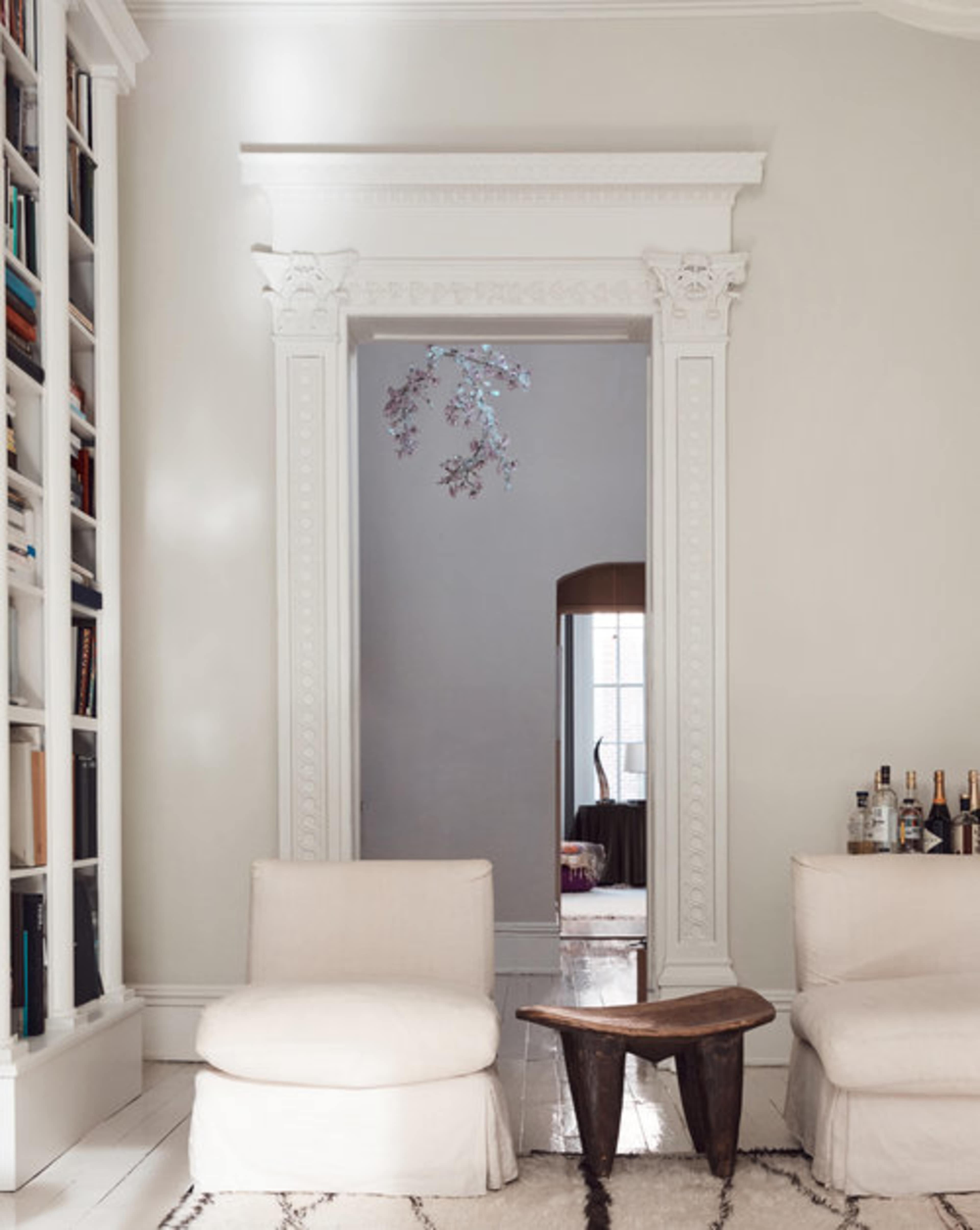 A minimalist living room features two white chairs, a wooden table, and a doorway with ornate trim leading to another room.