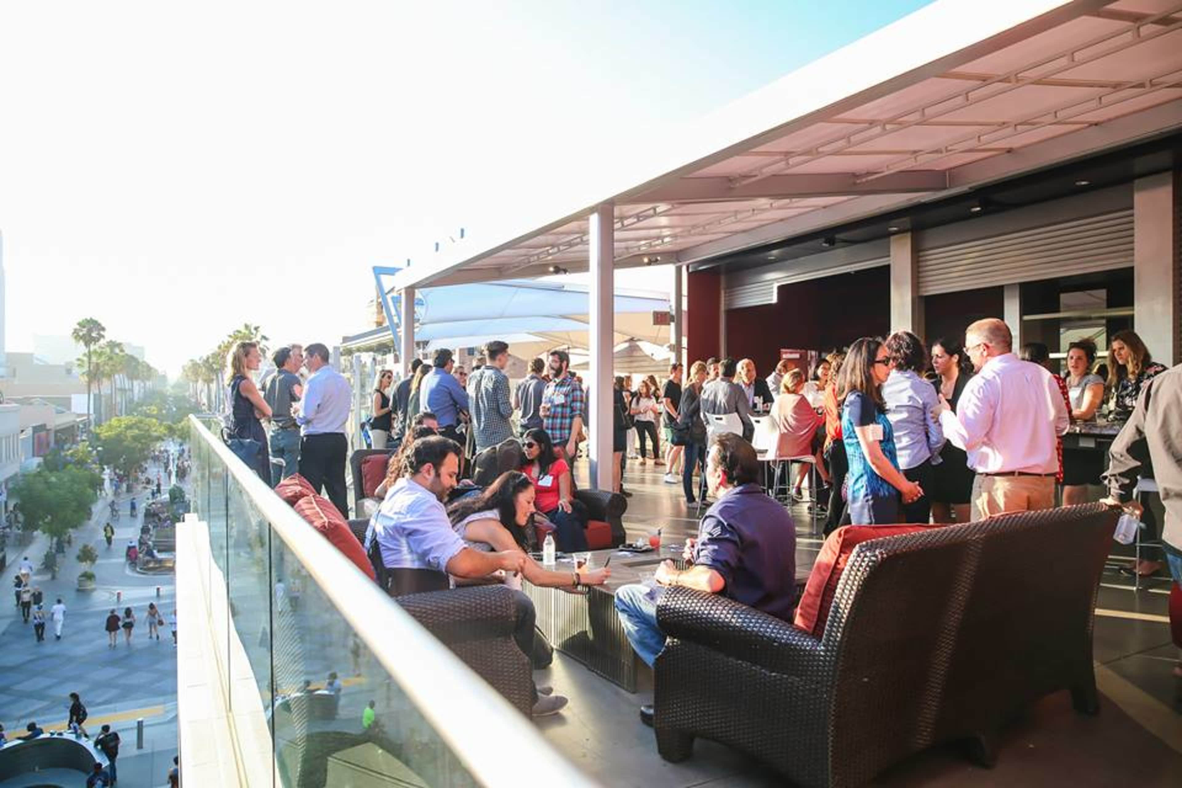 A group of people socializes on a rooftop terrace, enjoying a sunny day with a view of a pedestrian street below.
