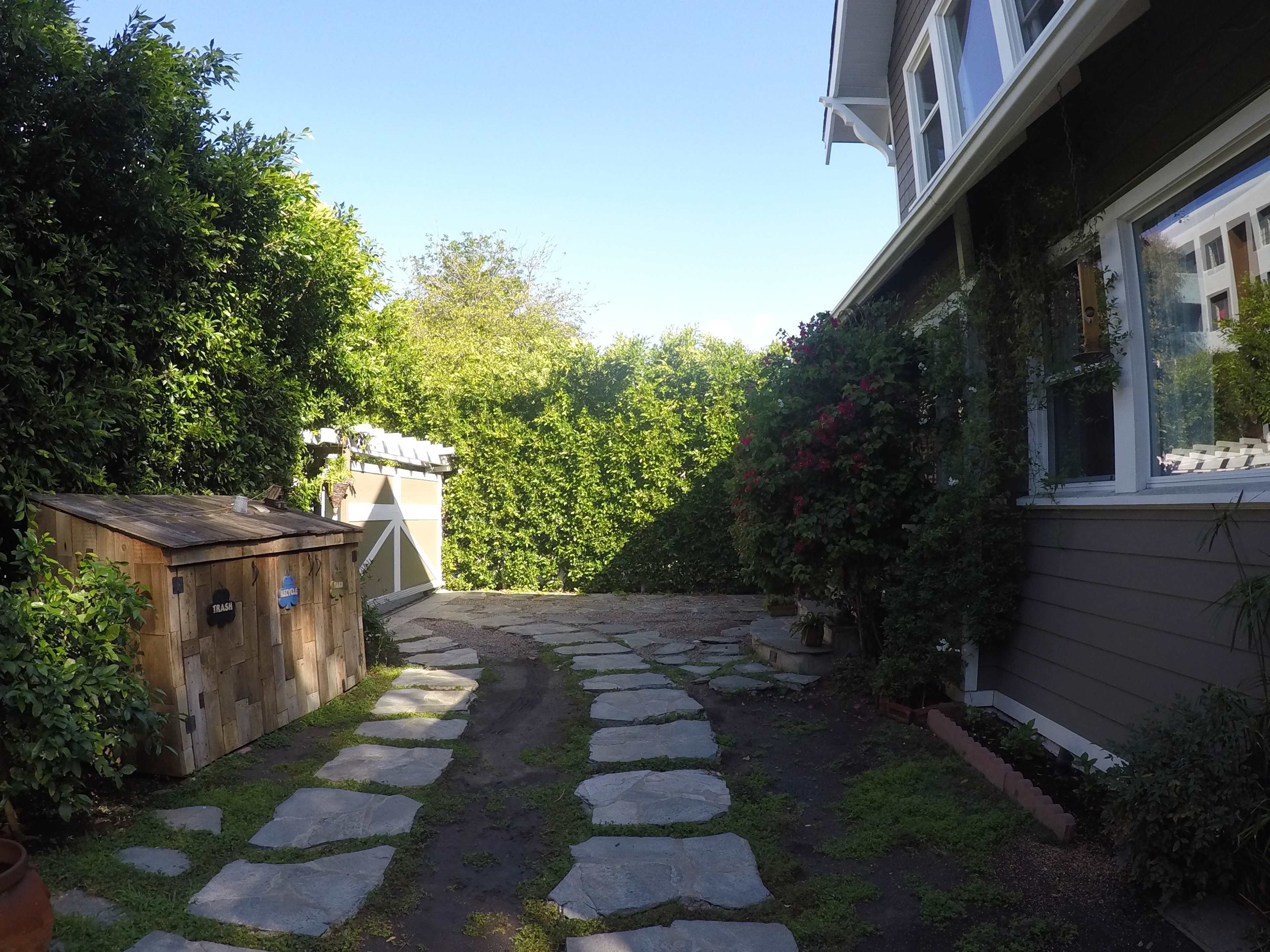 A stone pathway leads through a green yard beside a wooden shed and a house.