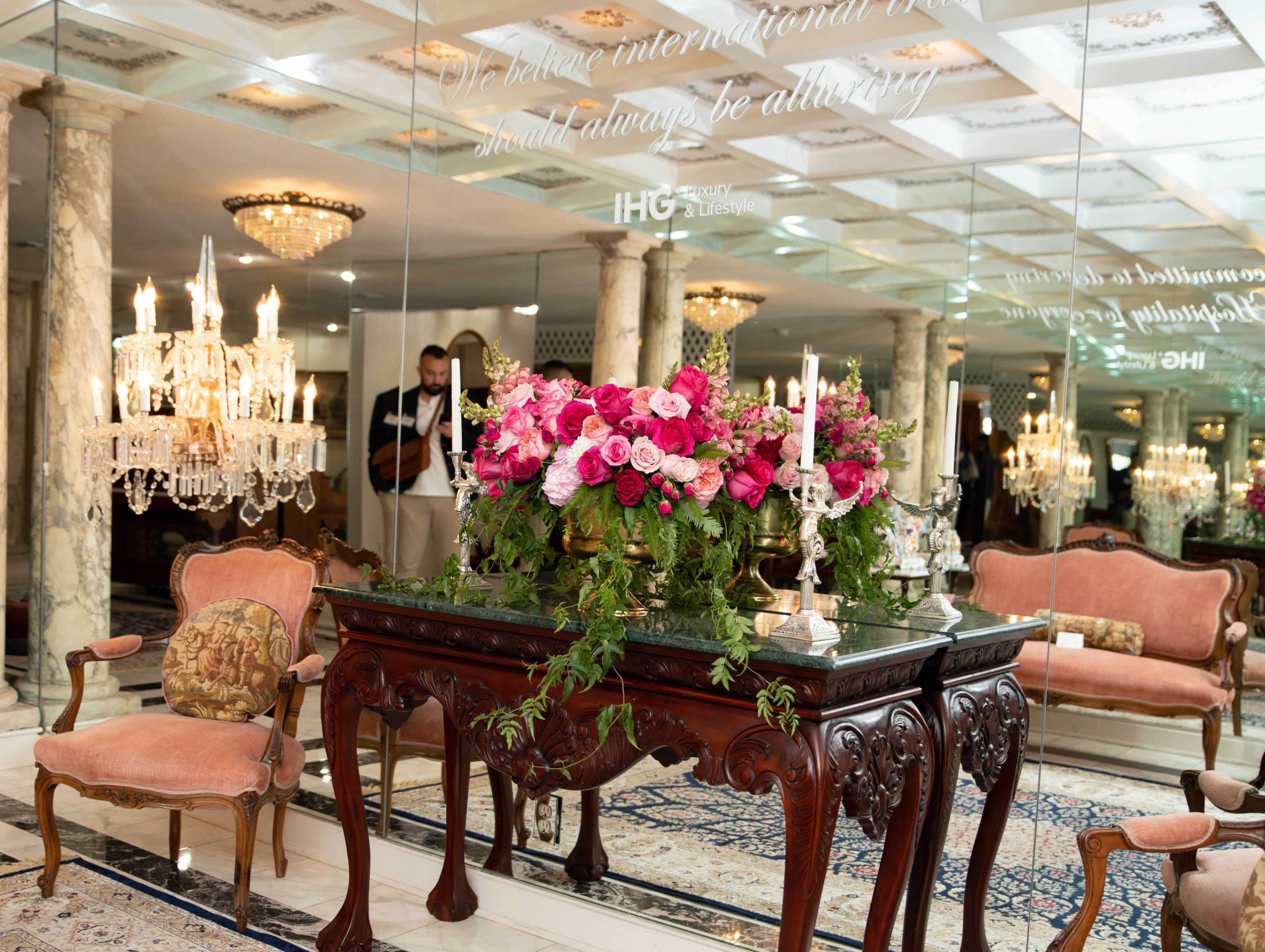 A wooden table adorned with a vibrant floral arrangement sits in front of a mirror in an elegantly decorated room illuminated by chandeliers.