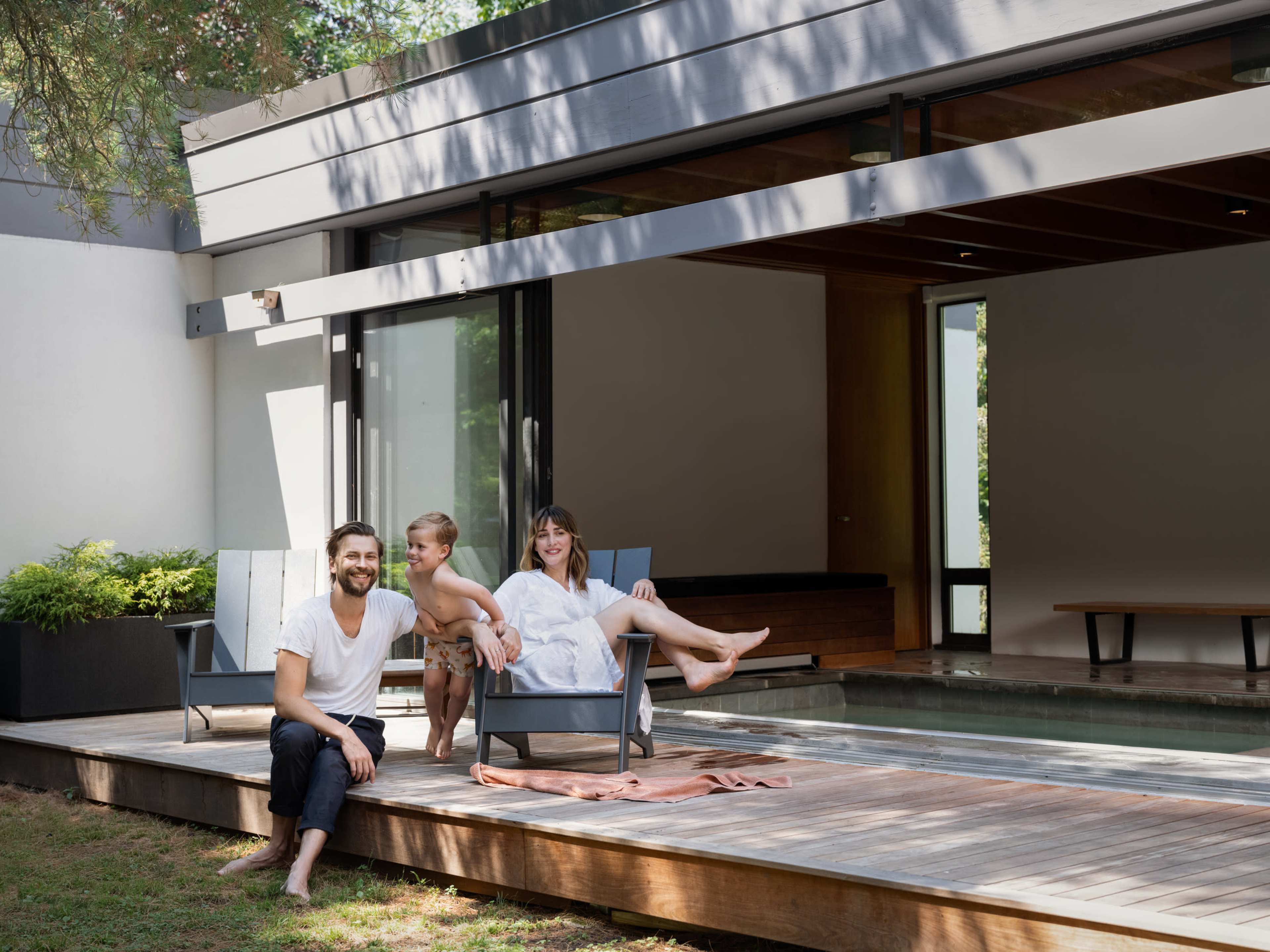 A man, a boy, and a woman sit on a wooden deck next to a pool in a modern home environment.