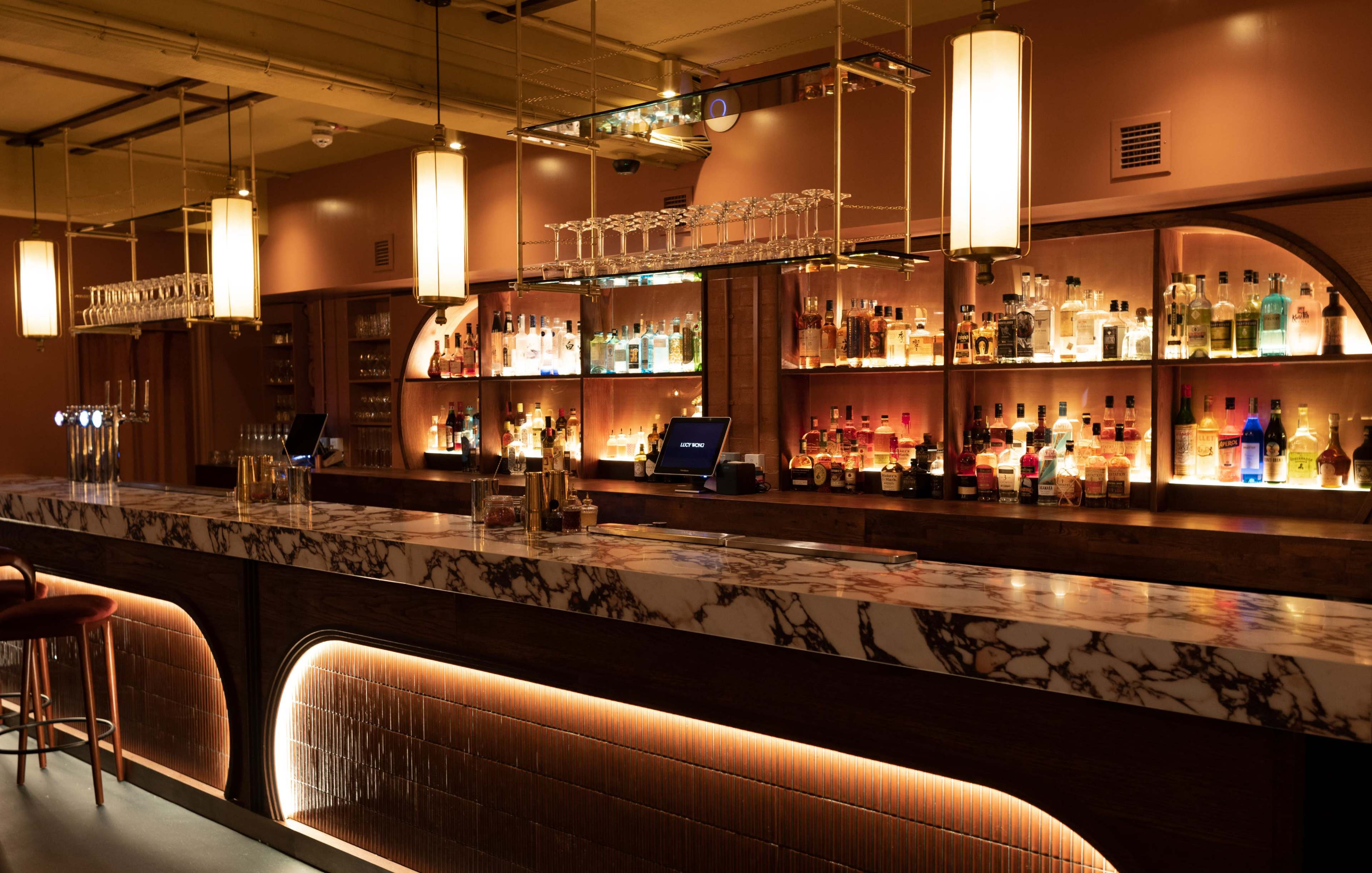 The image shows a stylish bar interior with a marble-topped counter, illuminated shelves filled with bottles, and pendant lighting.