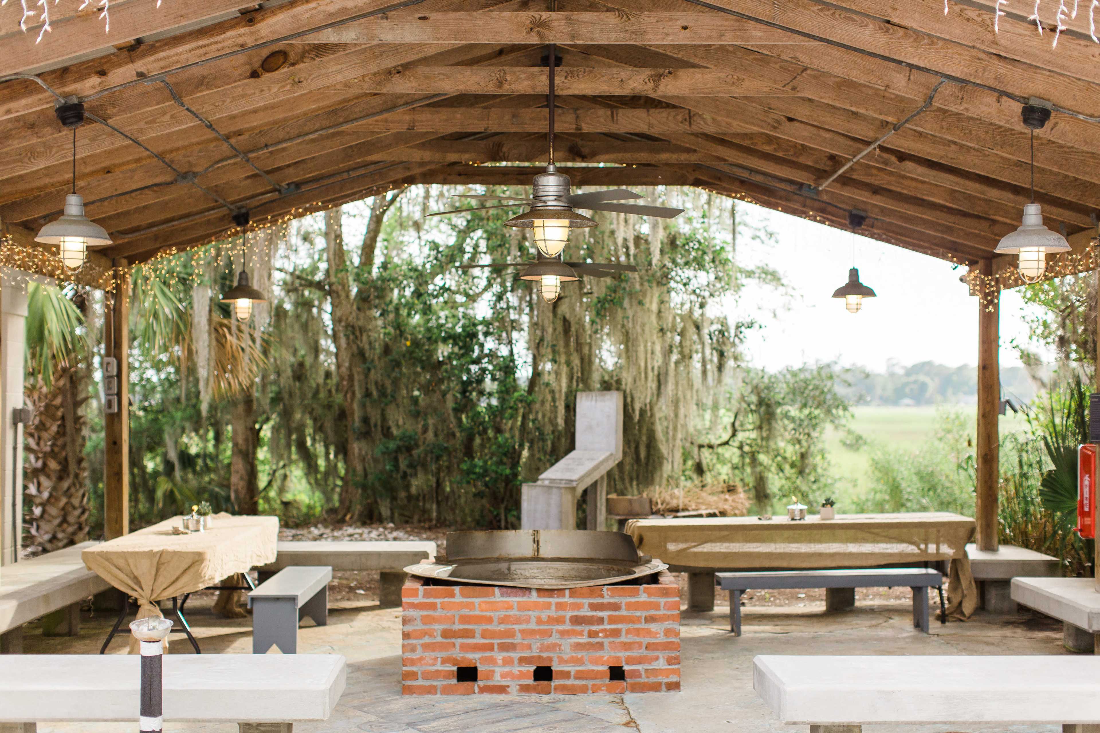 A covered outdoor gathering space with a brick fire pit in the center, surrounded by several tables and benches.