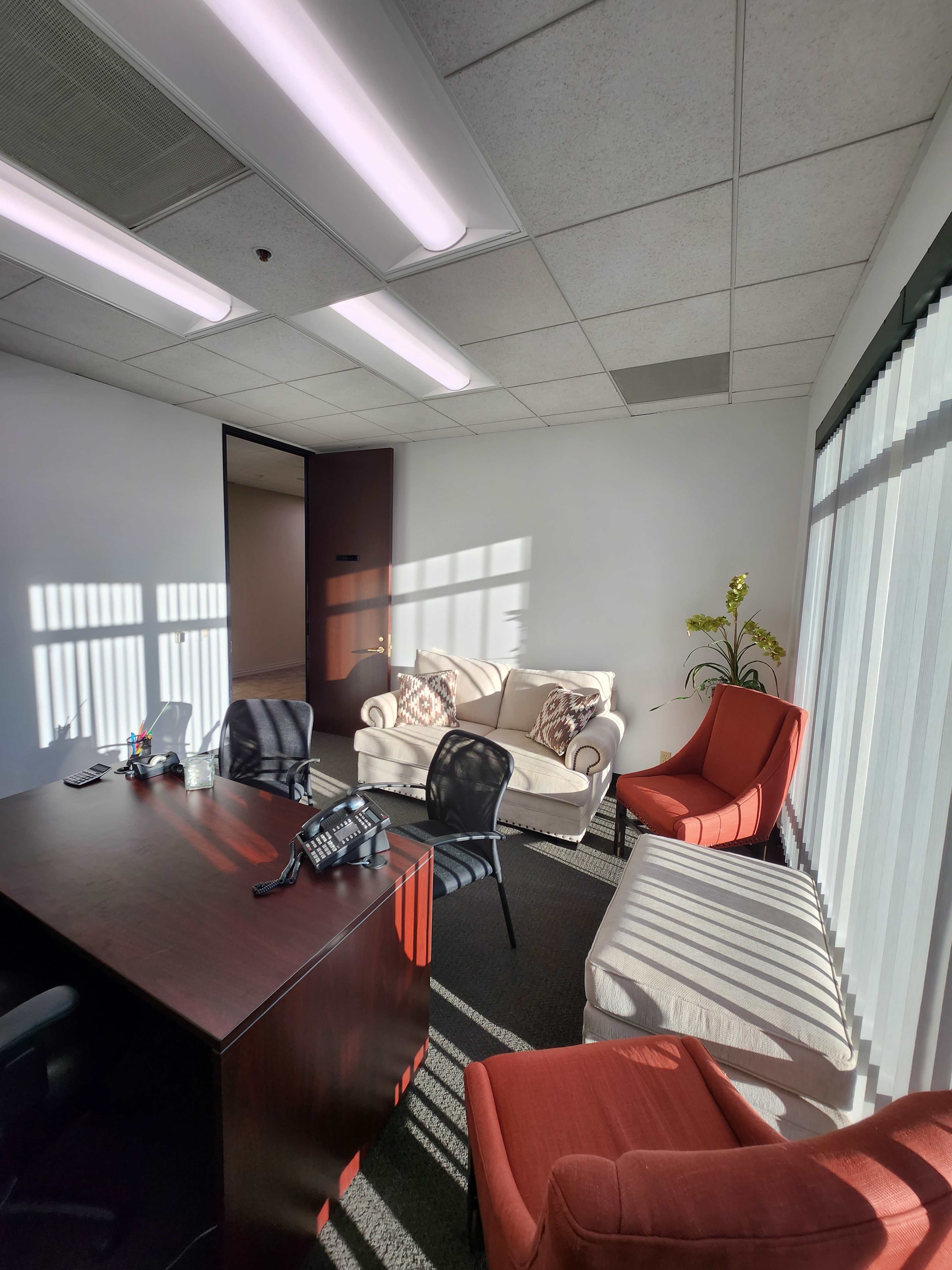 An office space features a wooden desk, a black telephone, and several seating options including two armchairs and a couch, illuminated by sunlight through the windows.
