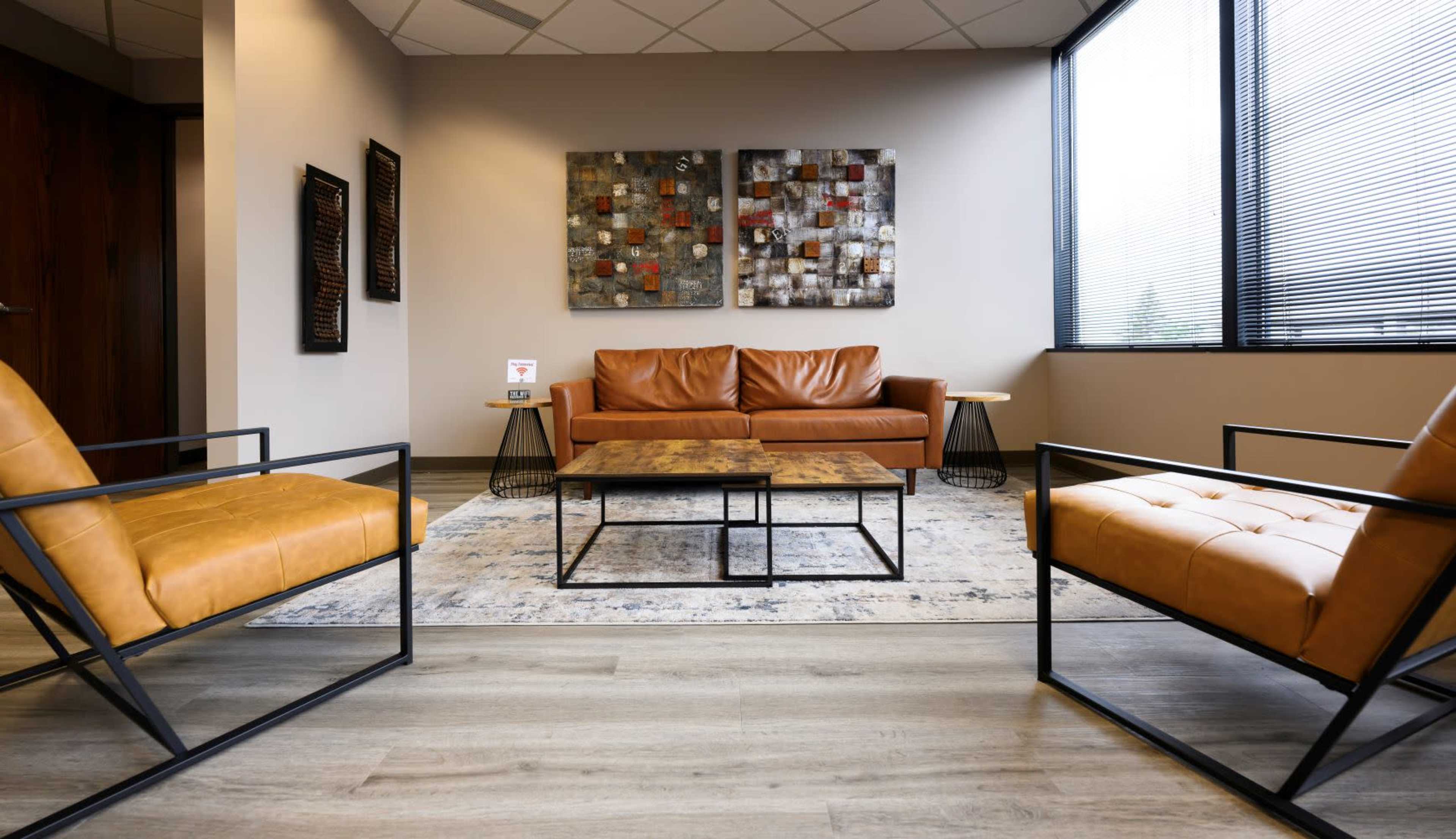 The image shows a modern seating area with a brown leather couch and two matching armchairs, a coffee table in the center, and abstract artwork on the walls.