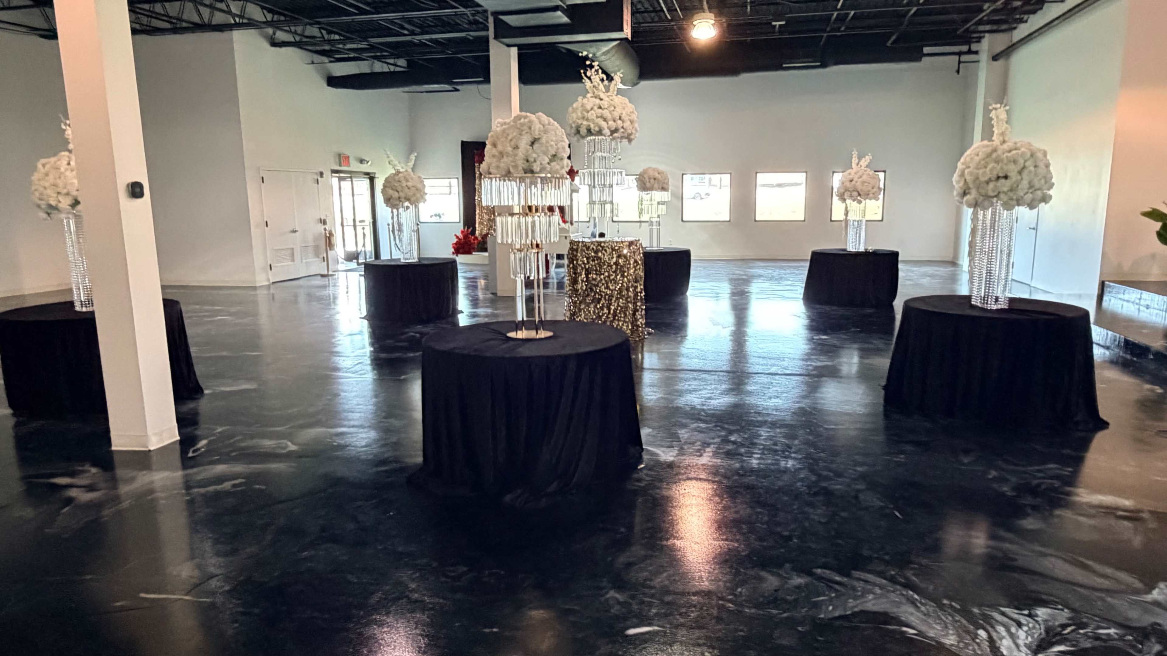The image shows an empty, spacious event venue featuring several tables covered with black cloth, each adorned with floral arrangements and decorative elements.