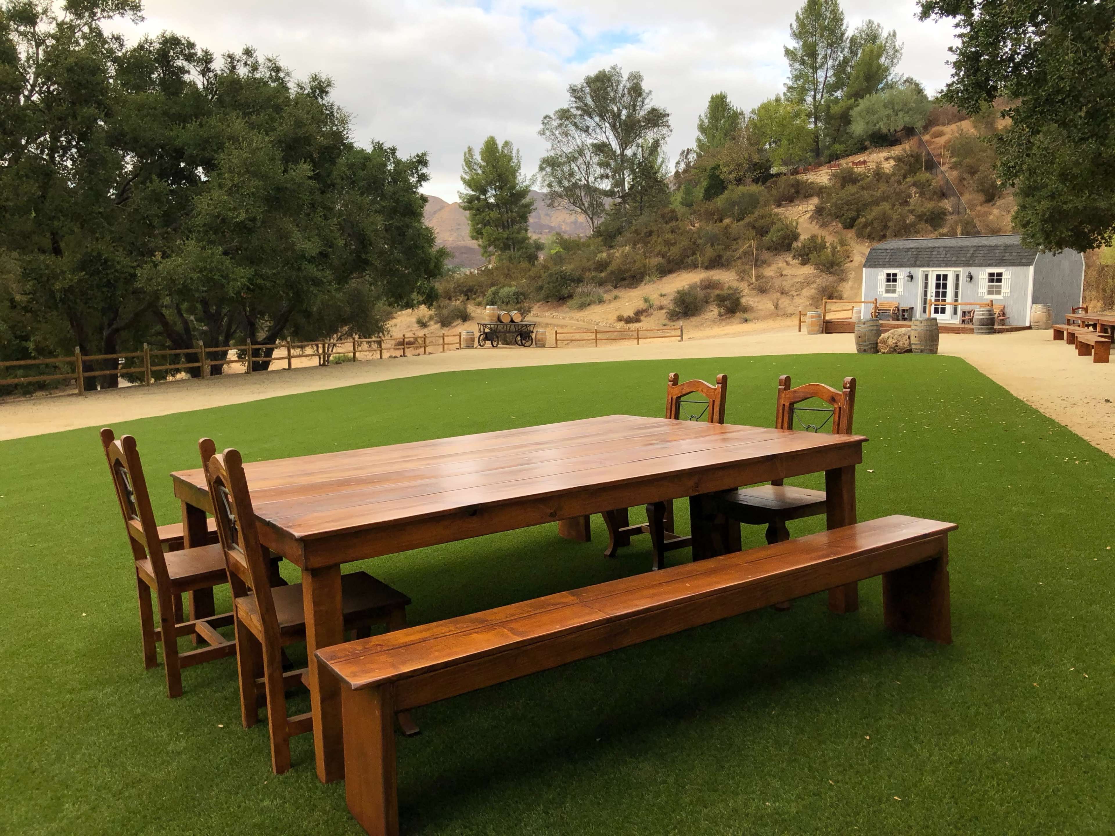 A large wooden table surrounded by chairs and a bench is set on a grassy area, with trees and hills visible in the background.
