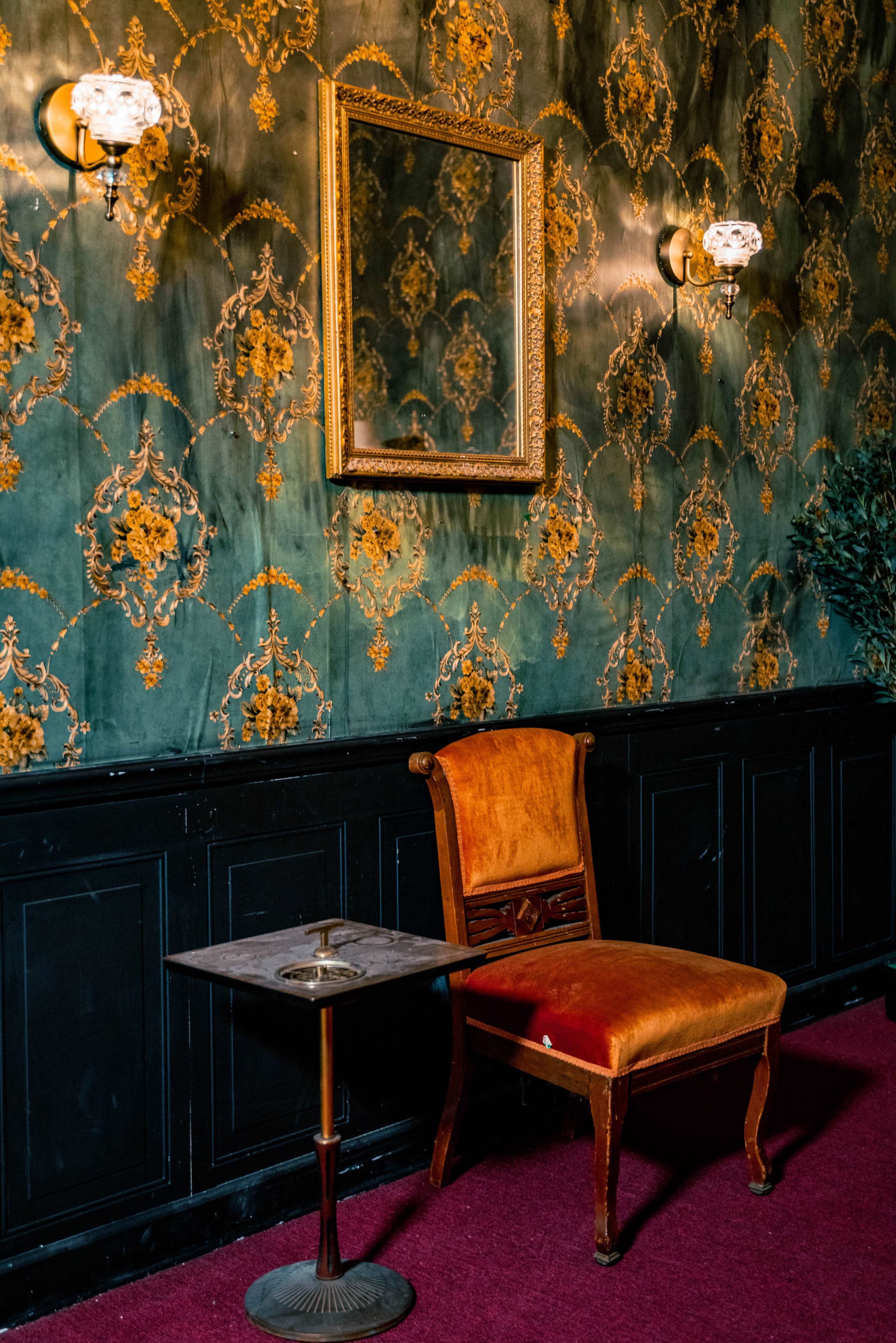 A corner of a room featuring a green and gold patterned wallpaper, an empty orange upholstered chair beside a small metal table with an ashtray, and a framed mirror on the wall.