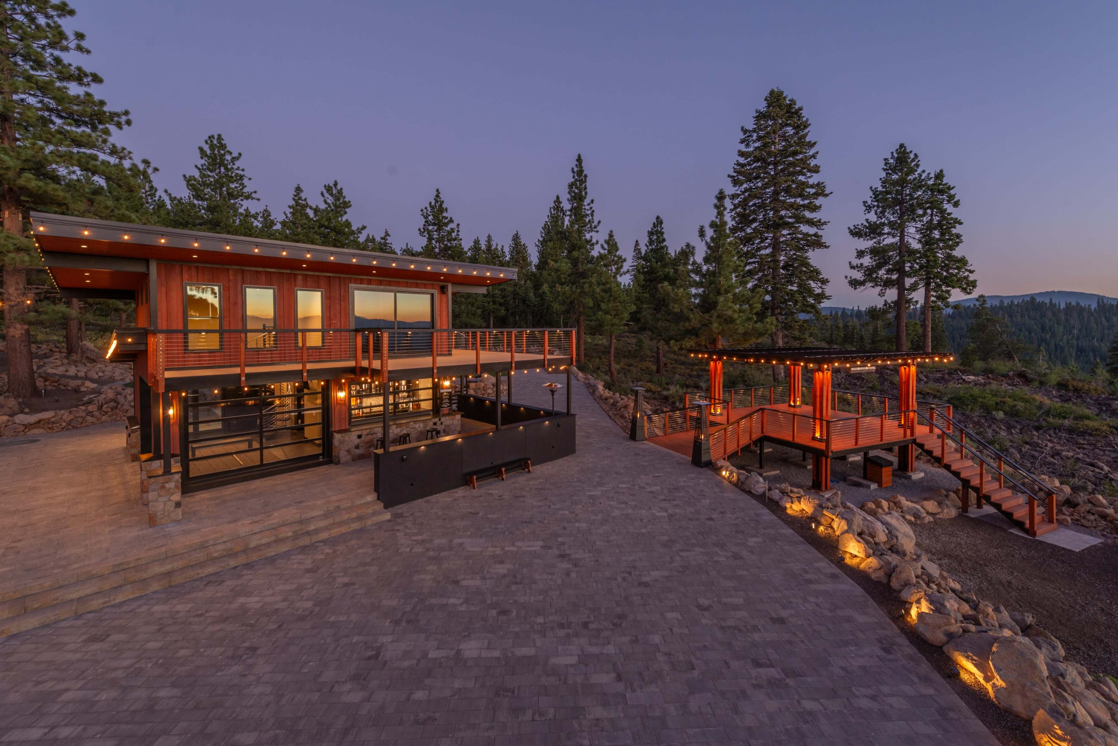 The image shows a modern cabin with a wooden deck, surrounded by tall trees and a patio area illuminated by outdoor lights.