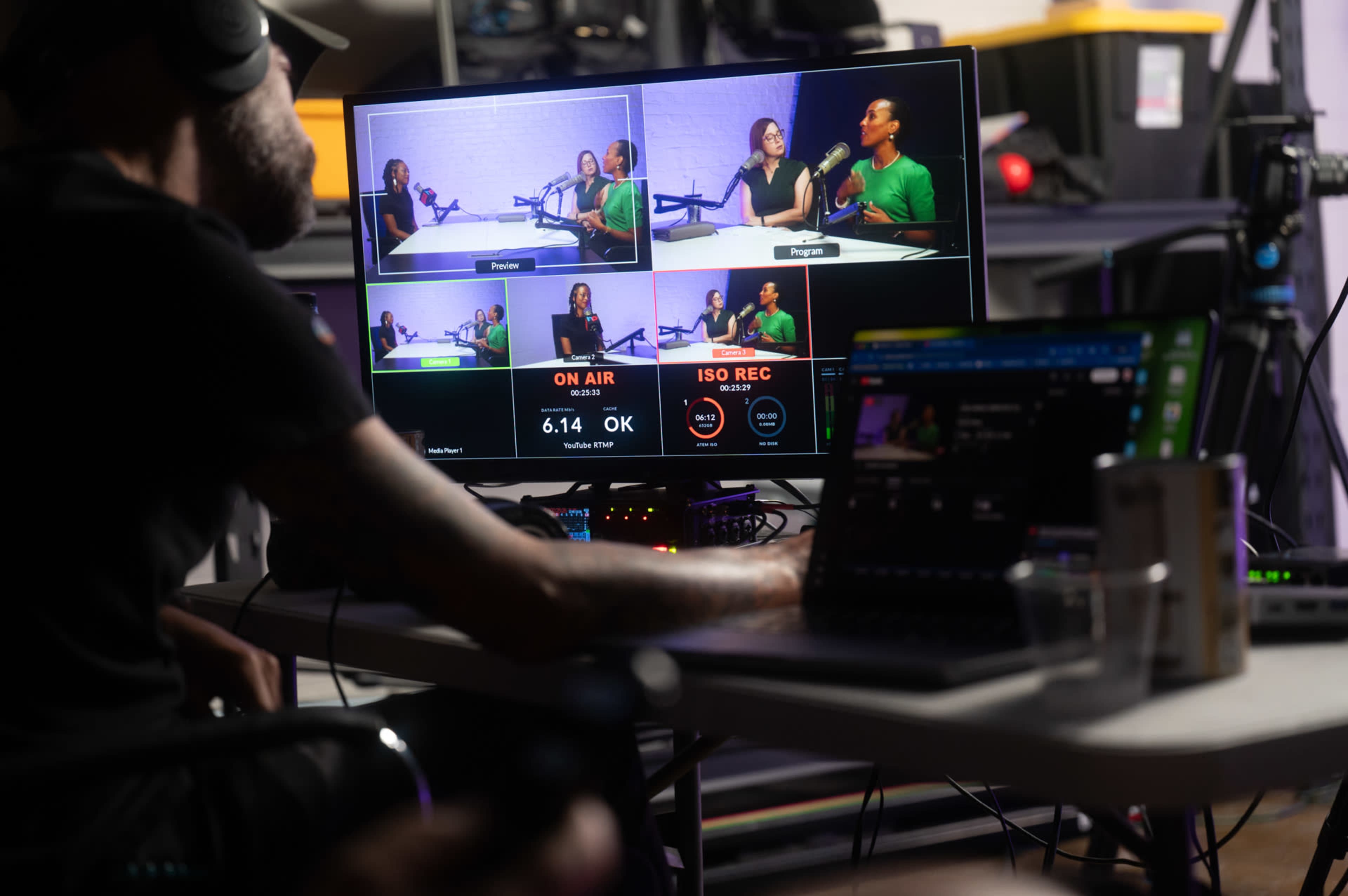 A person is operating a video production setup with multiple screens displaying live feeds from a podcast recording session.