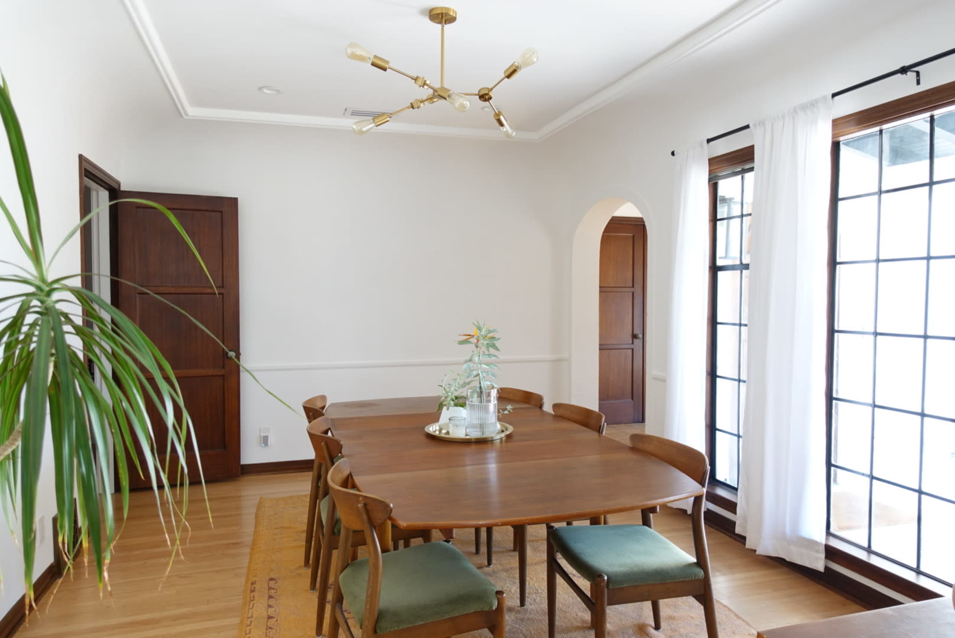 The image shows a bright dining room with a wooden table surrounded by chairs, a ceiling light fixture, and large windows featuring white curtains.