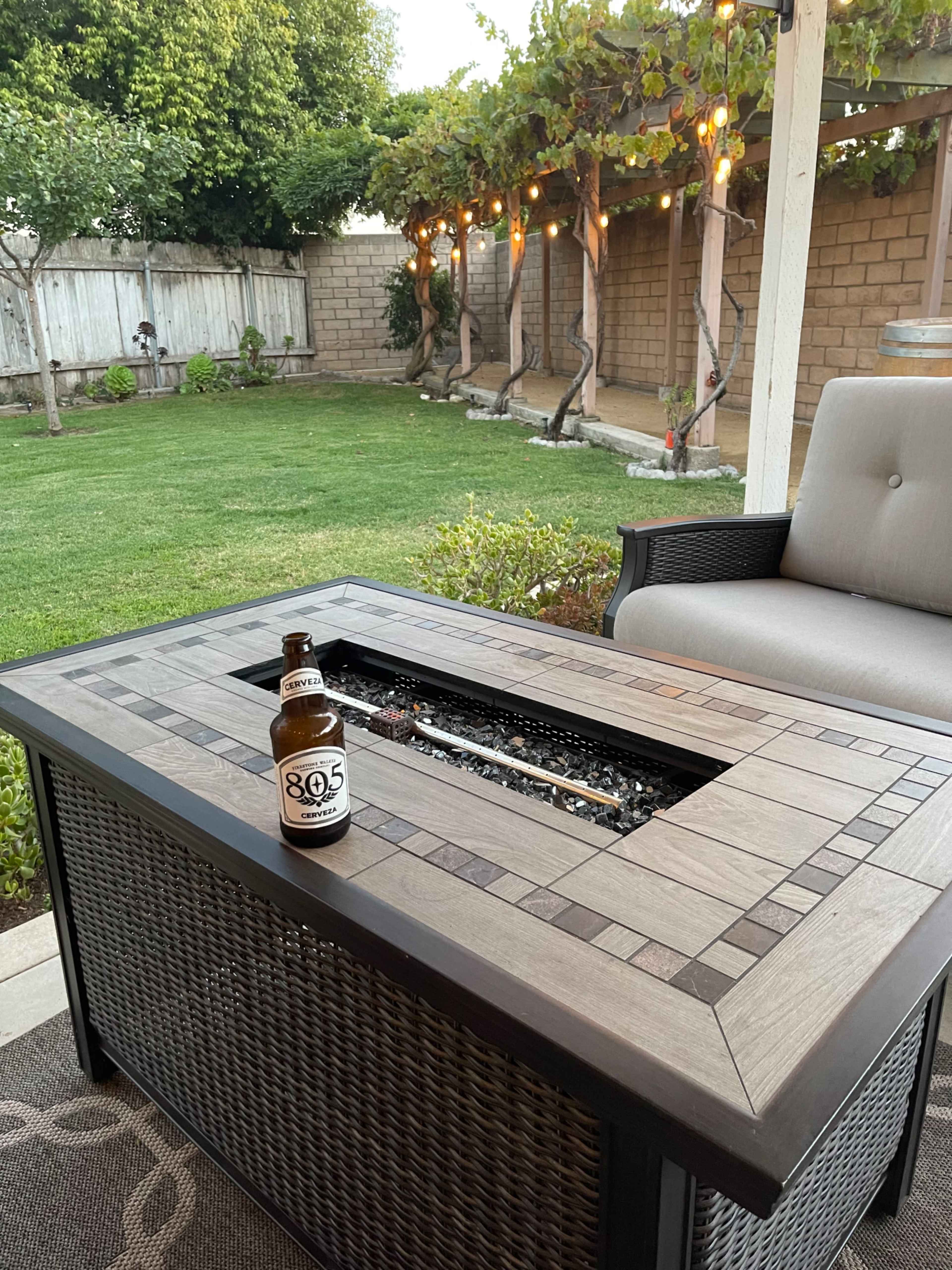 A backyard patio features a fire table and a bottle of beer on top, surrounded by well-maintained greenery and string lights.