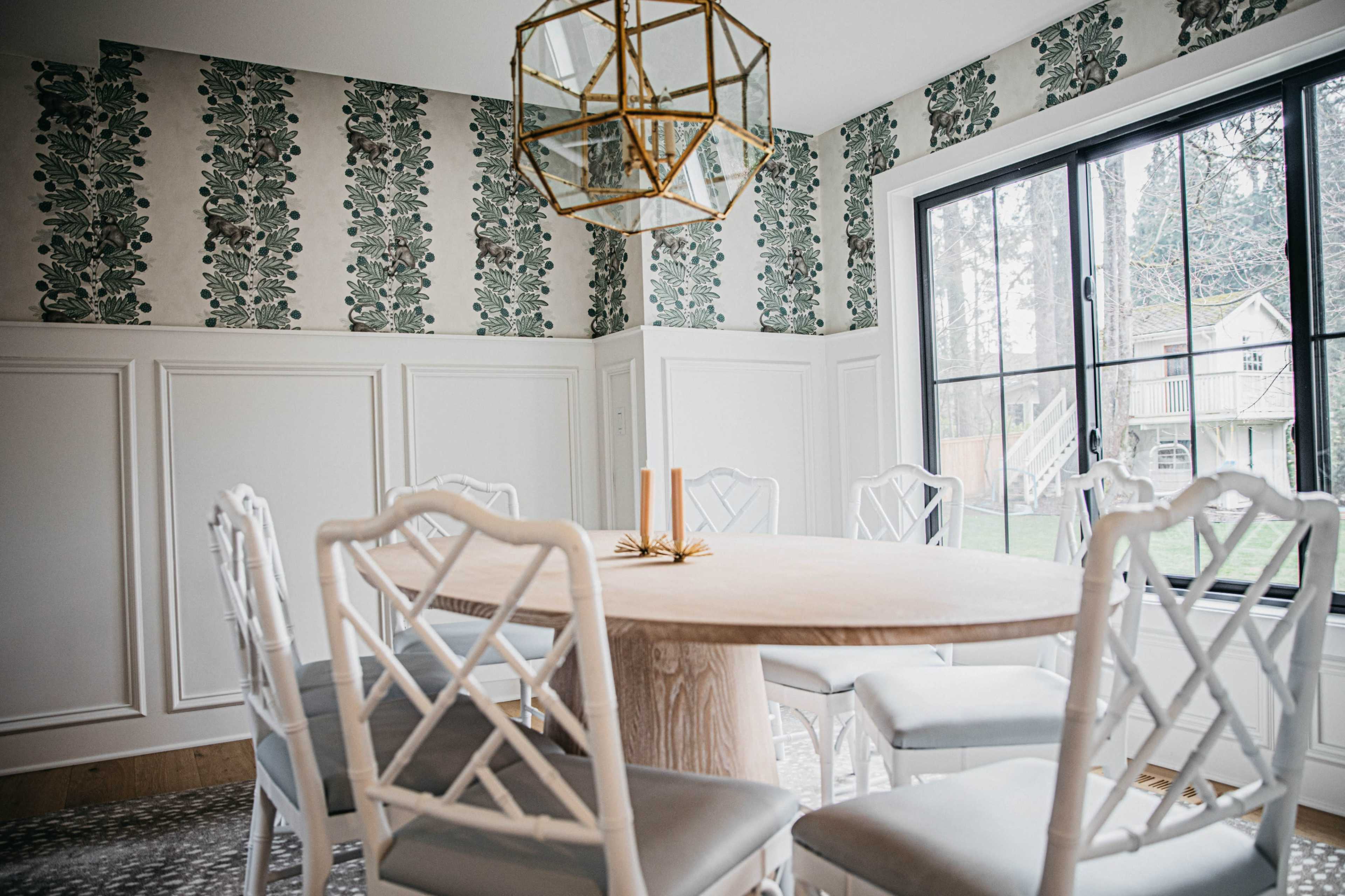 A round wooden dining table is surrounded by six white chairs in a room with floral wallpaper and a large window letting in natural light.
