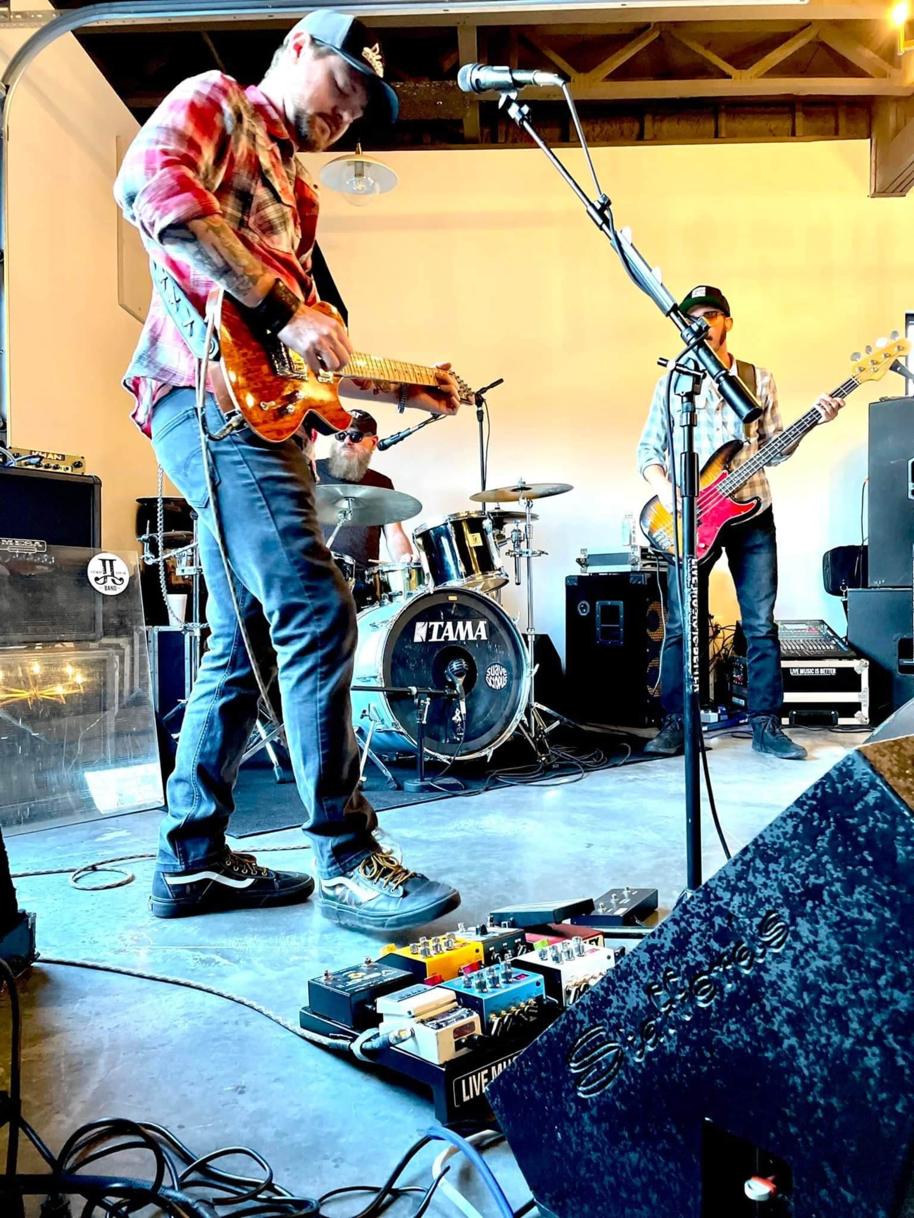 Three musicians perform in a rehearsal space, with one guitarist adjusting effects pedals while the drummer and bassist play behind him.