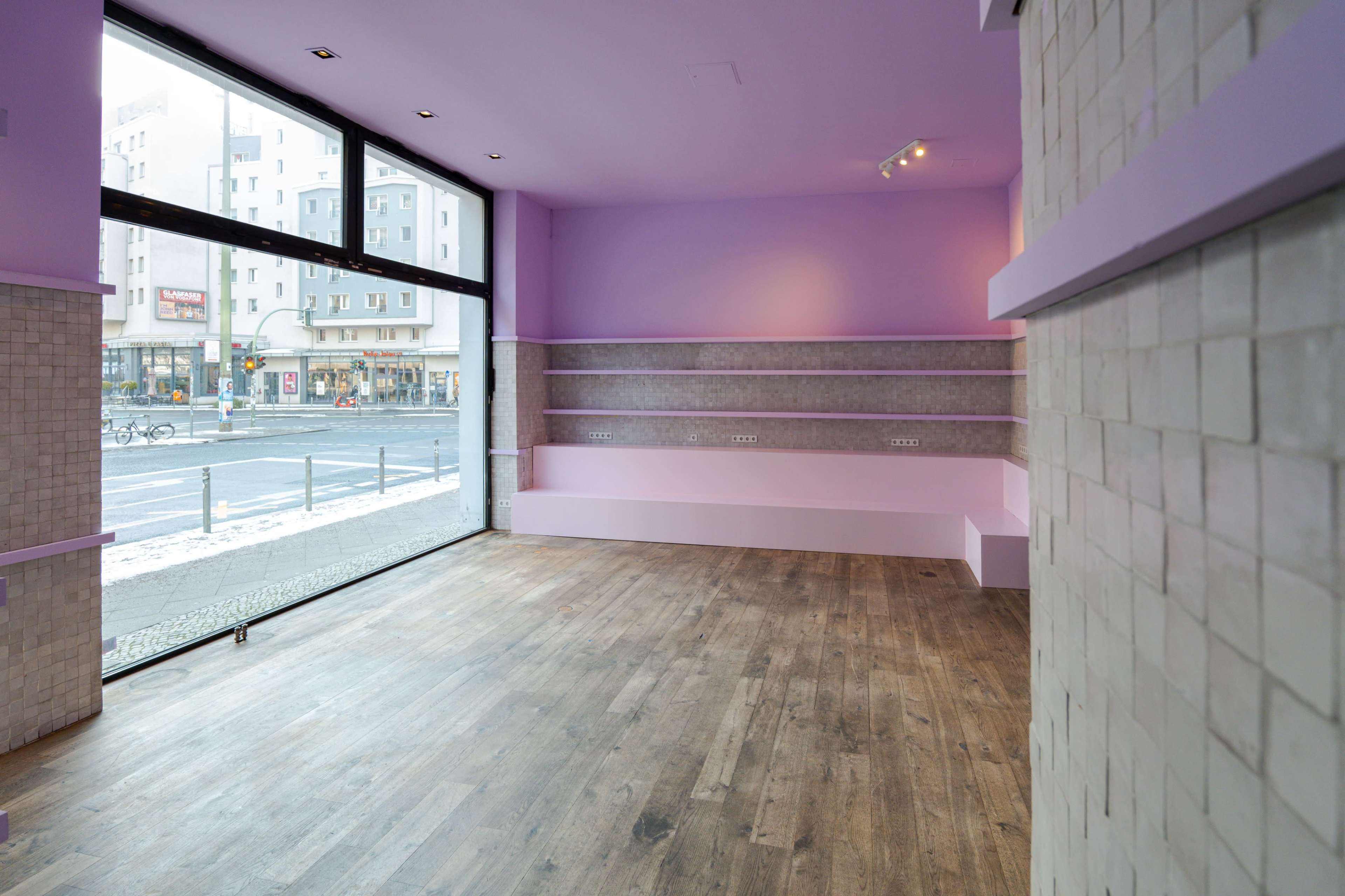 An empty retail space with purple walls and wooden flooring, featuring large windows that provide a view of the street outside.