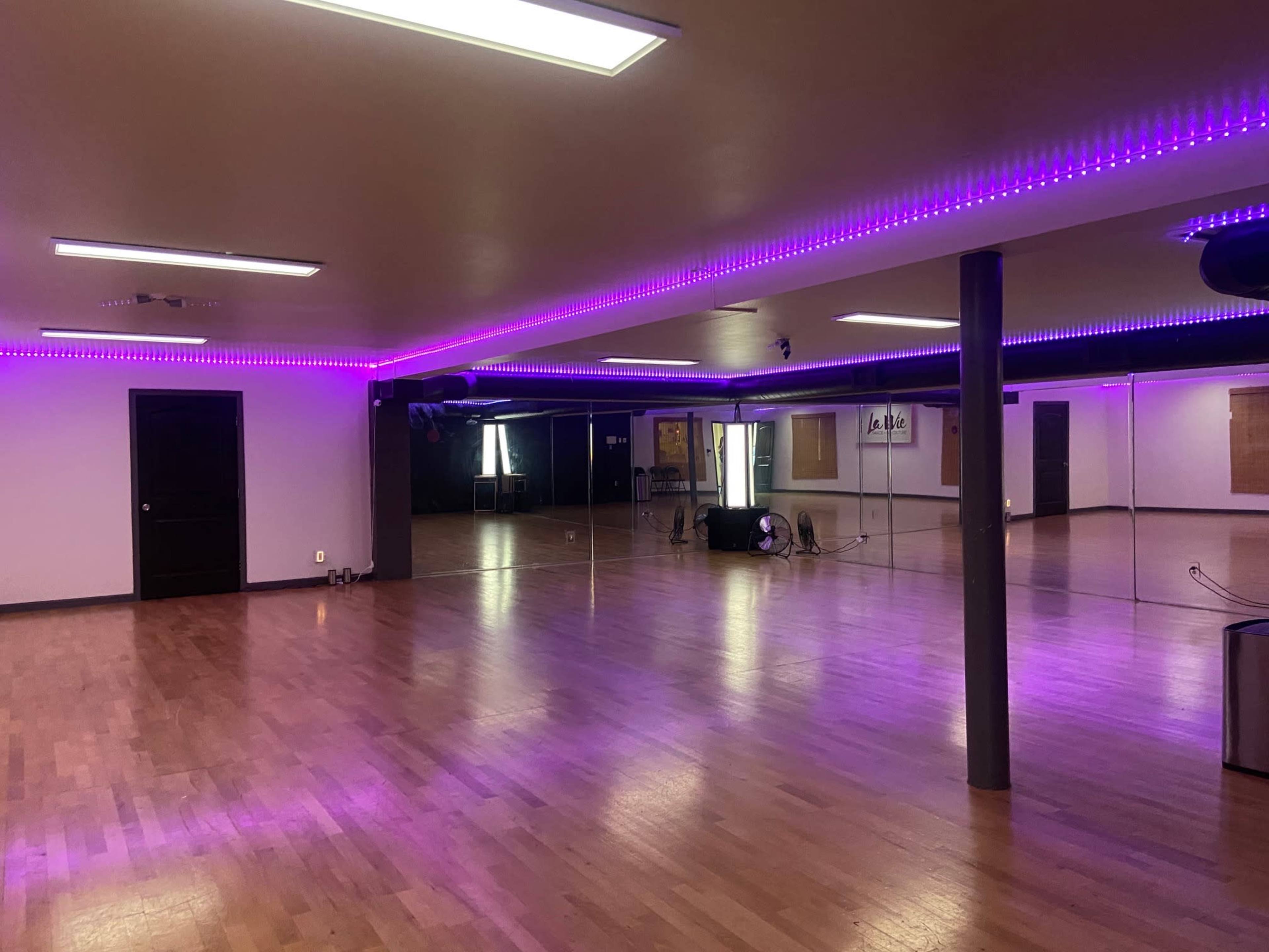 A spacious dance studio features mirrored walls, wooden floors, and purple LED lighting along the ceiling.