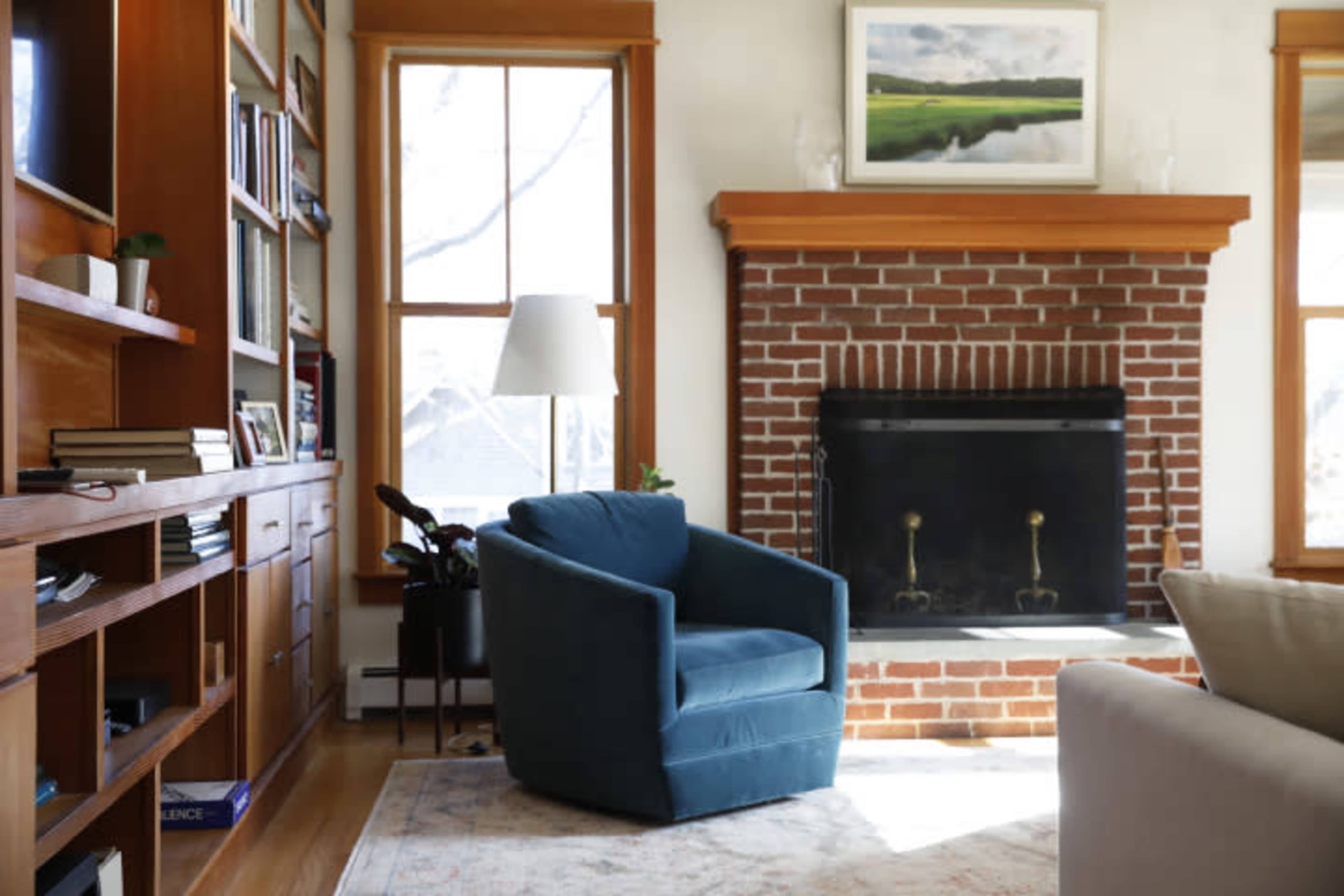 The image shows a cozy living room with a blue armchair facing a brick fireplace and shelves filled with books and decorative items.