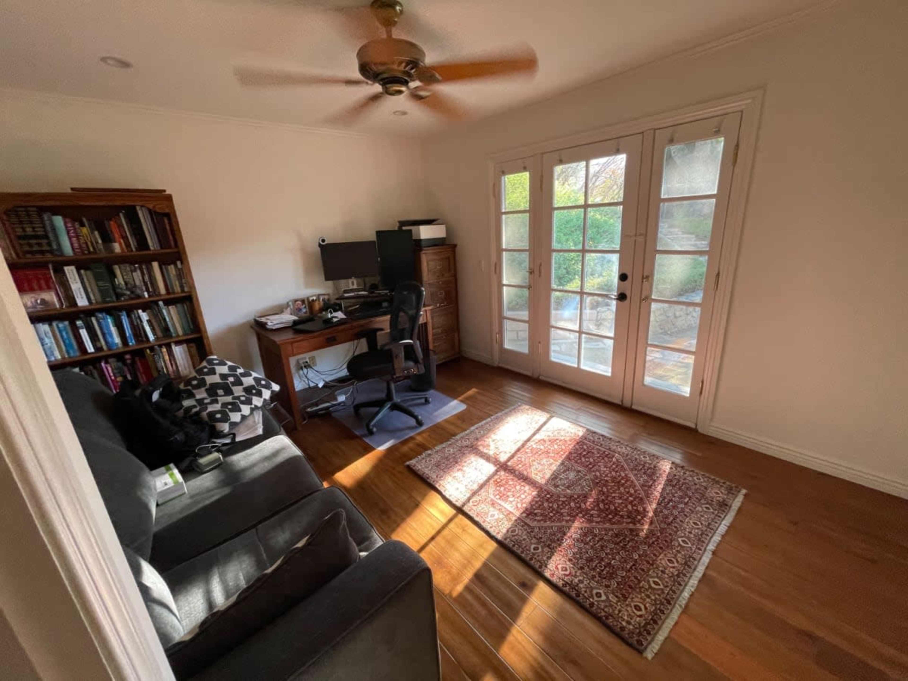 A small home office features a desk with a computer, a chair, a bookshelf, and large French doors that lead to an outdoor area.