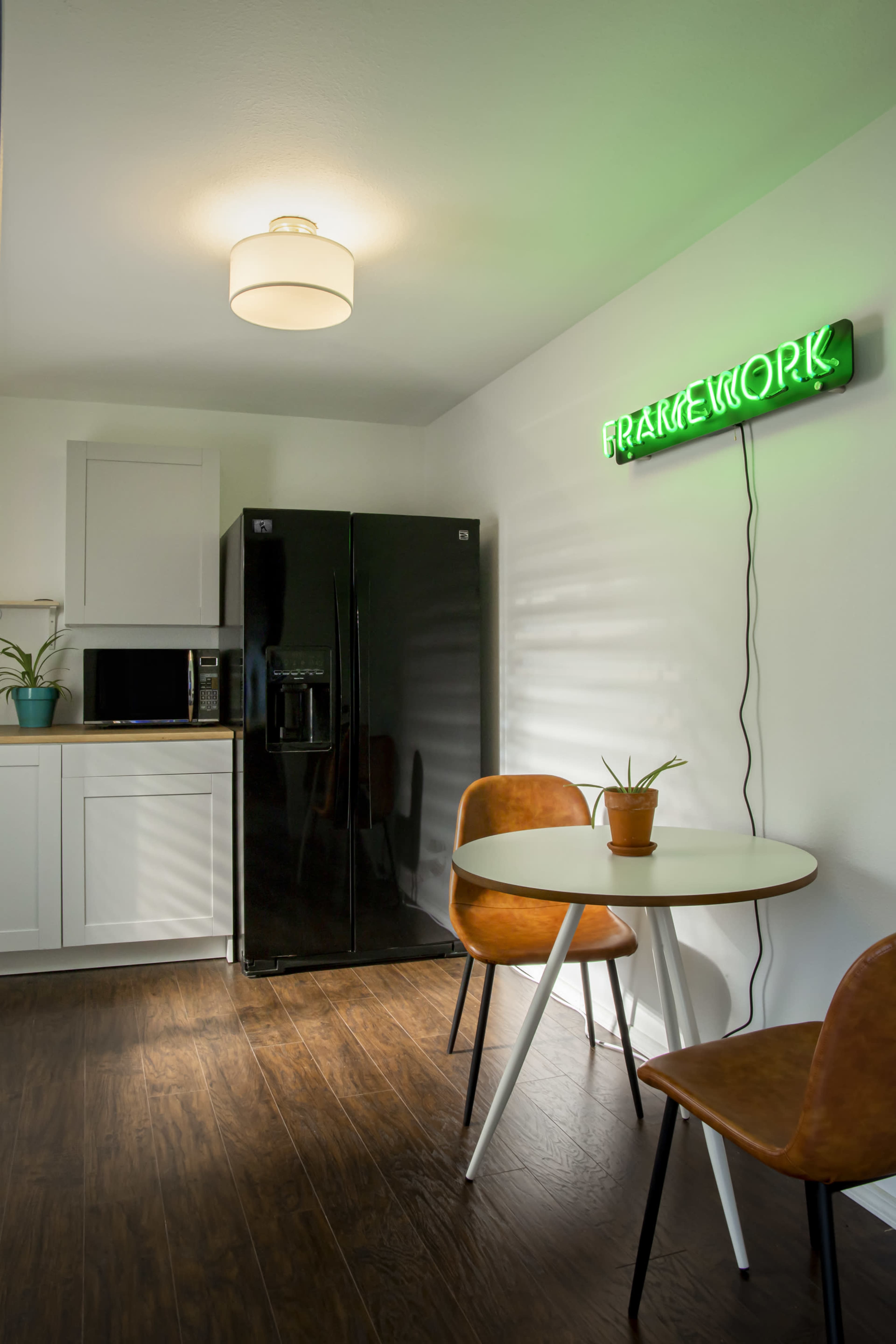 A modern kitchen features a black refrigerator, a microwave, a small table with two chairs, and a neon sign on the wall.