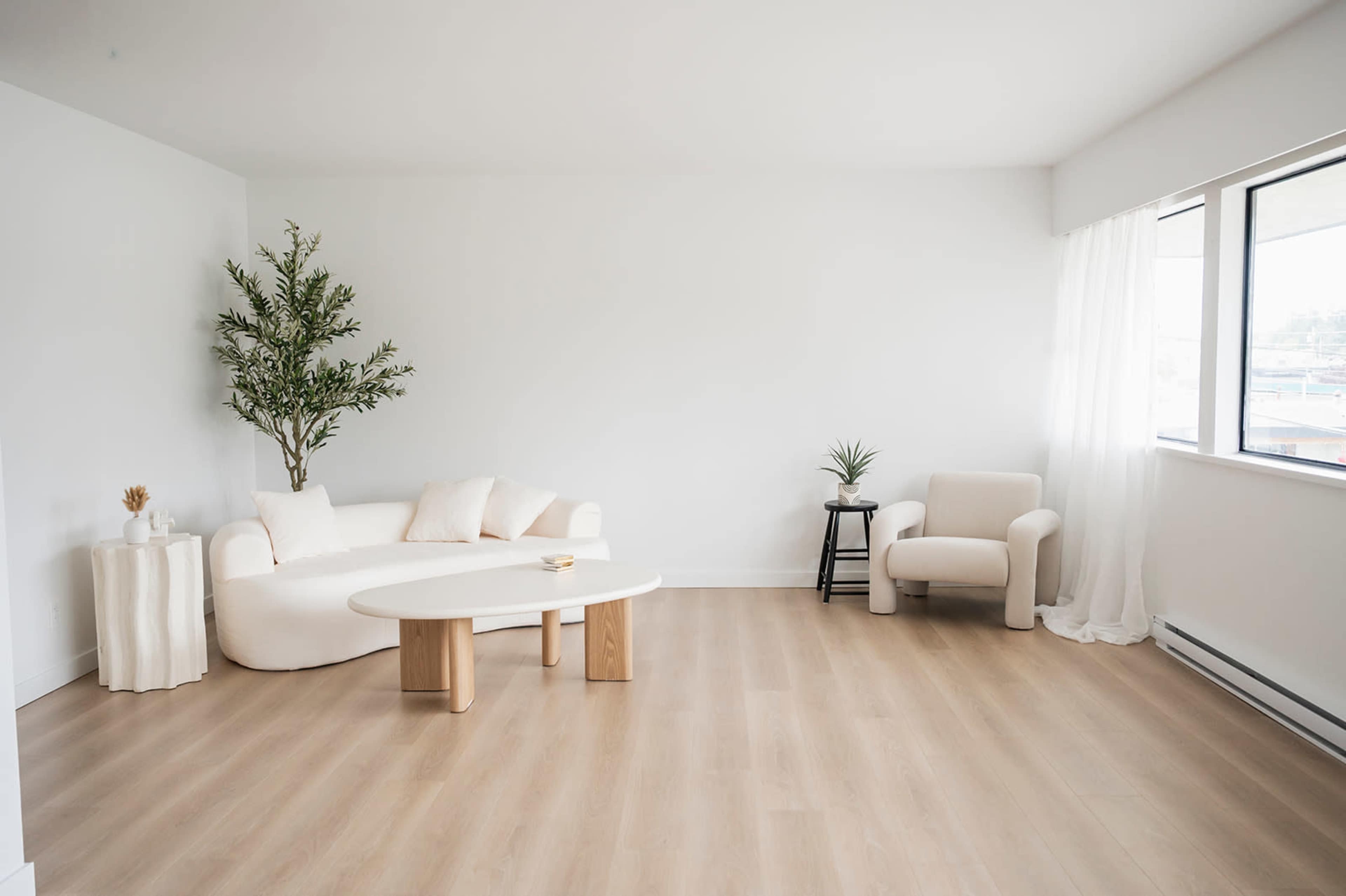 The image shows a minimalist living room featuring a curved white sofa, a round coffee table, and a light-colored armchair, along with a plant and a small side table.