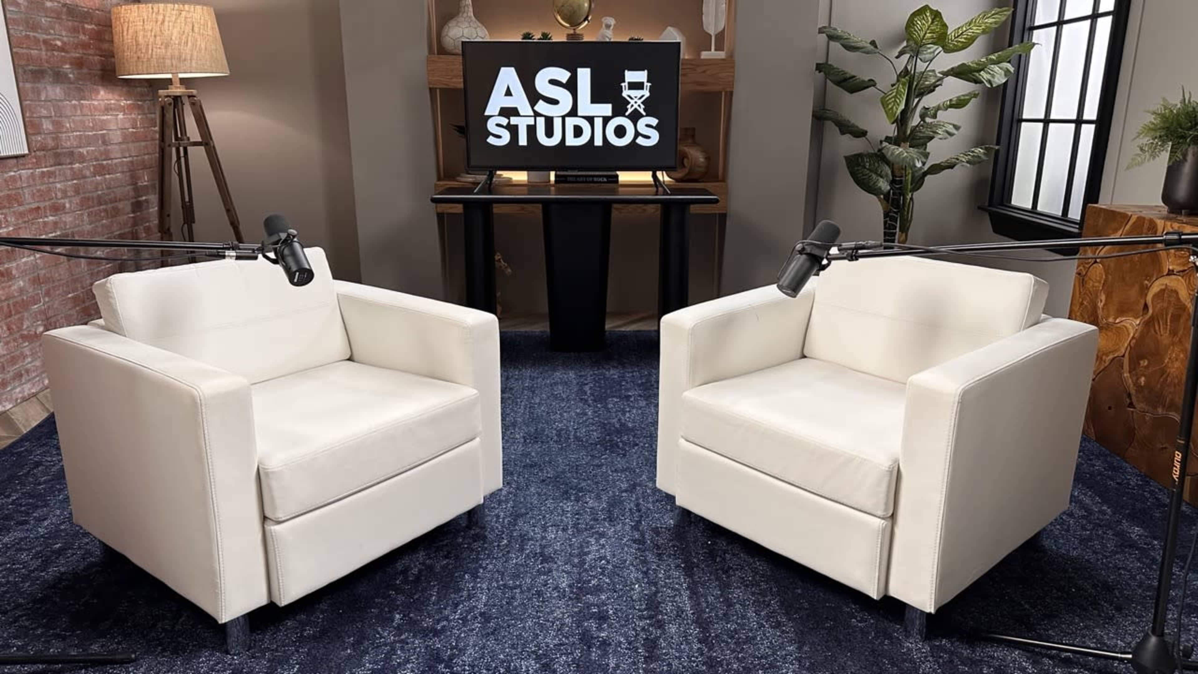 The image shows two white armchairs with microphones in front of a television displaying the logo "ASL STUDIOS," set in a well-lit studio environment.