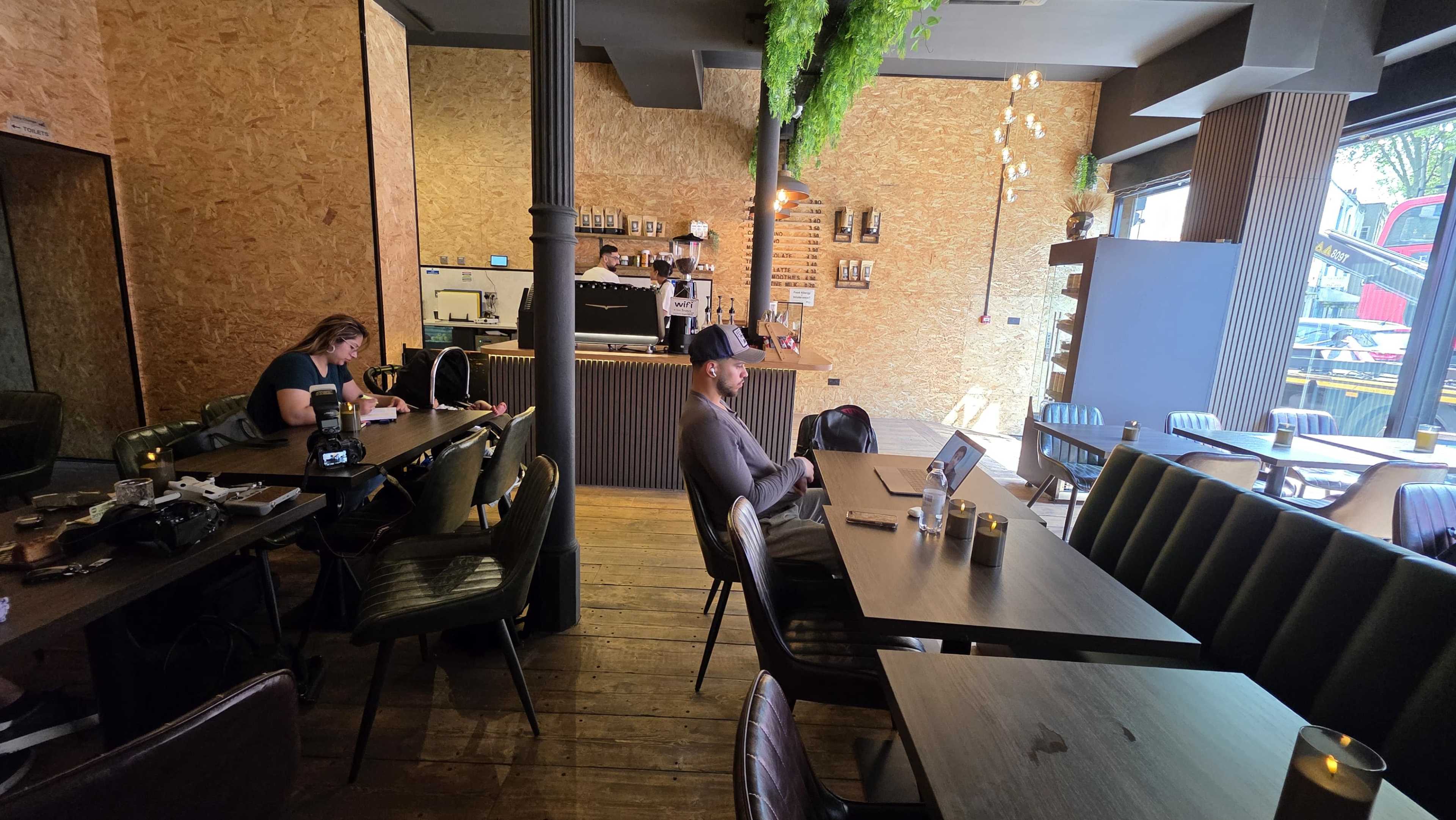 A café interior features wooden flooring, tables, and people working on laptops with a large window showing an urban setting outside.