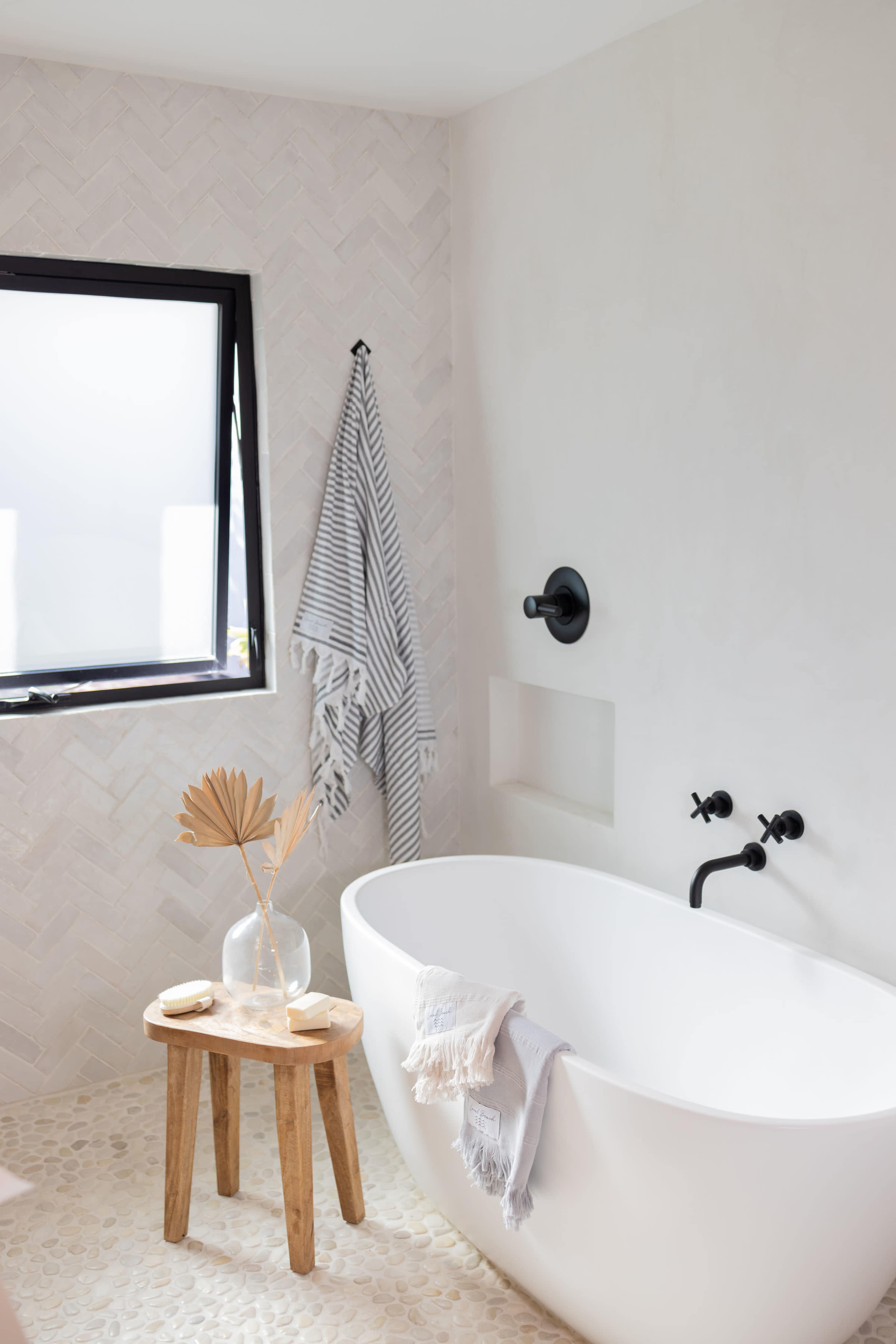 A minimalistic bathroom featuring a freestanding white bathtub, a wooden stool with a decorative vase and dried leaves, and a window with a monochrome striped towel hanging nearby.