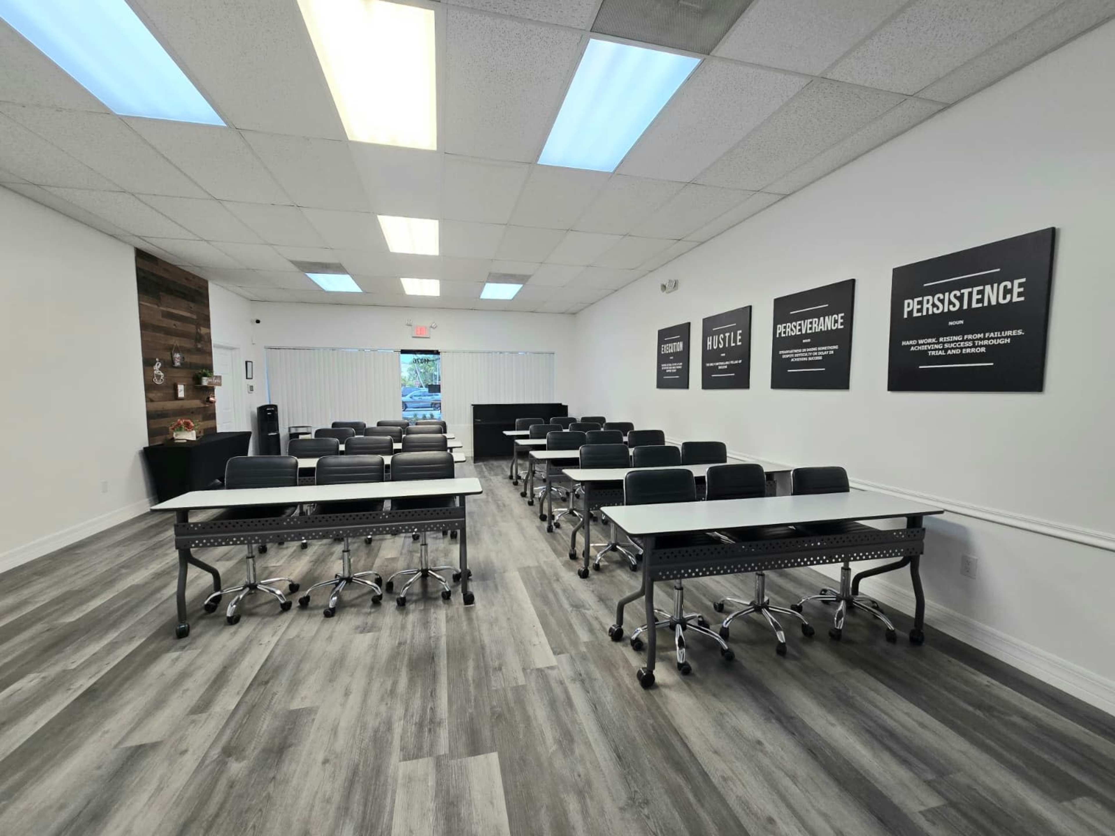 The image shows a neatly arranged classroom with several rows of tables and chairs, illuminated by overhead lights and large windows.