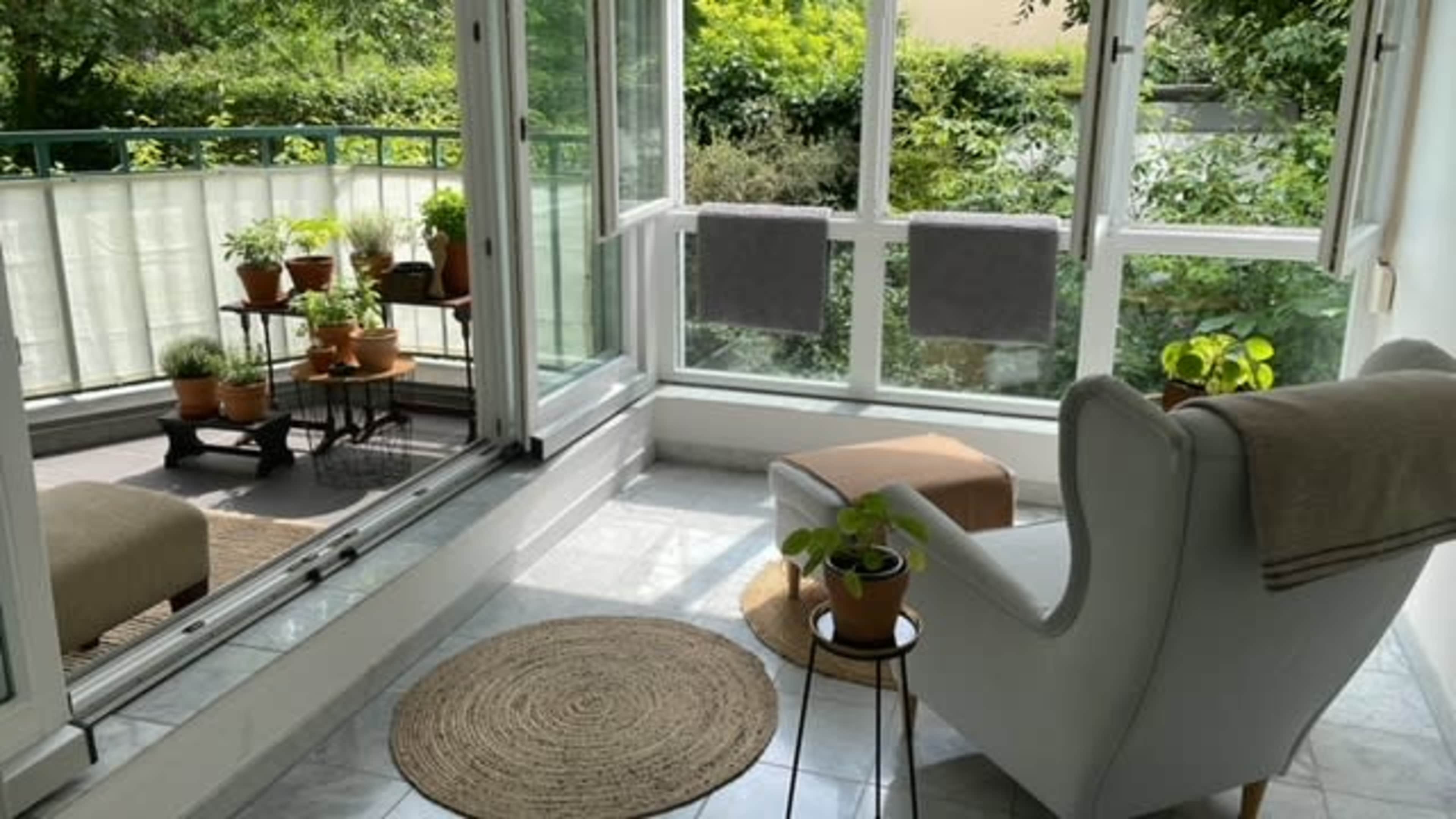 A sunlit sunroom features a cozy chair, a small table with a potted plant, and several plants on a metal cart, surrounded by large windows open to a green view.